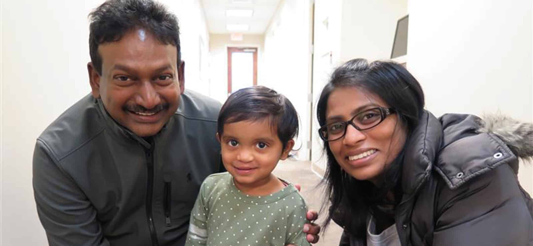 A child and two persons smile in a brightly lit hallway with white walls, creating a cheerful, familial atmosphere.