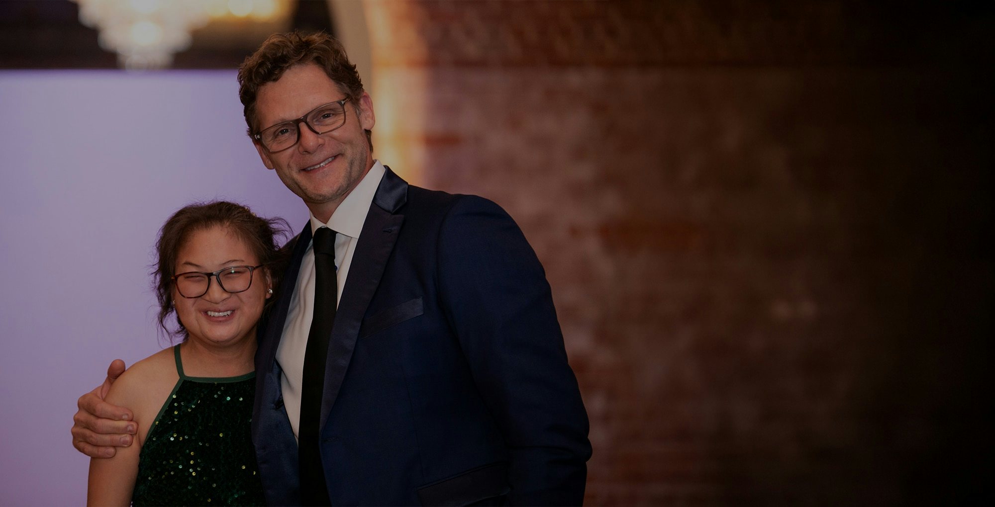A person in a suit hugs another in a green dress, posing indoors against a brick wall and a softly lit background.