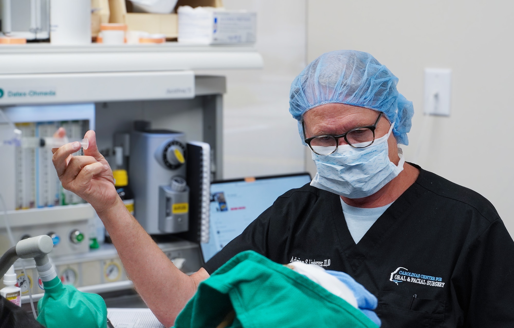 A person in medical scrubs and mask holds a syringe in a clinical setting, surrounded by medical equipment.