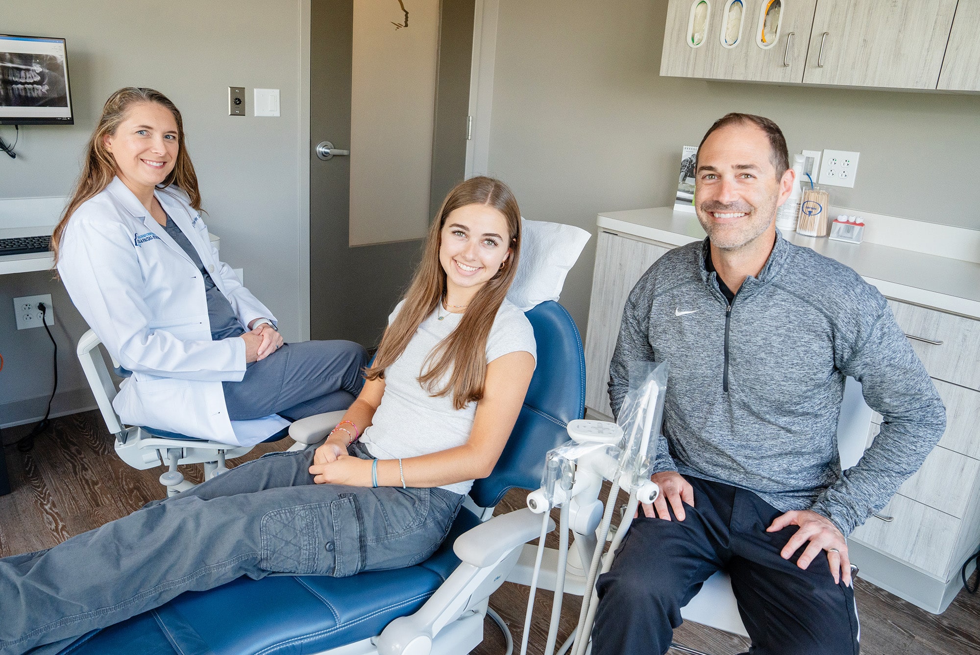 Three people in a dental office with equipment visible. One person is seated in a dentist chair, smiling.