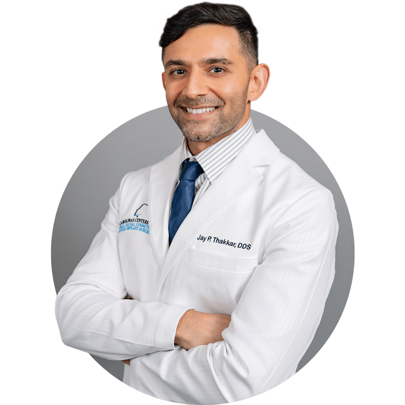 A person in a white coat with a blue tie smiles, standing against a gray background, signifying a professional healthcare setting.