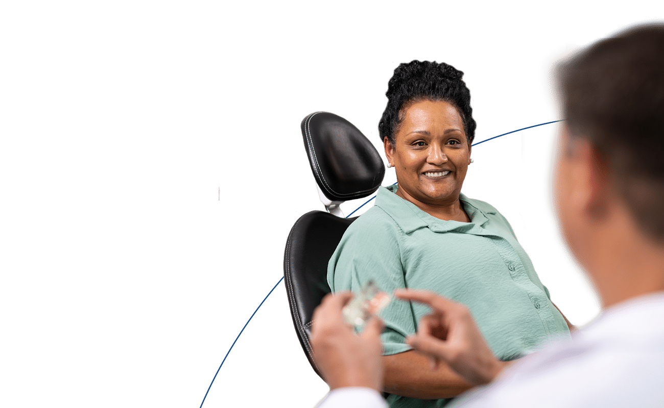 A person is smiling while seated in a chair, interacting with another person holding dental tools against a simple blue background.
