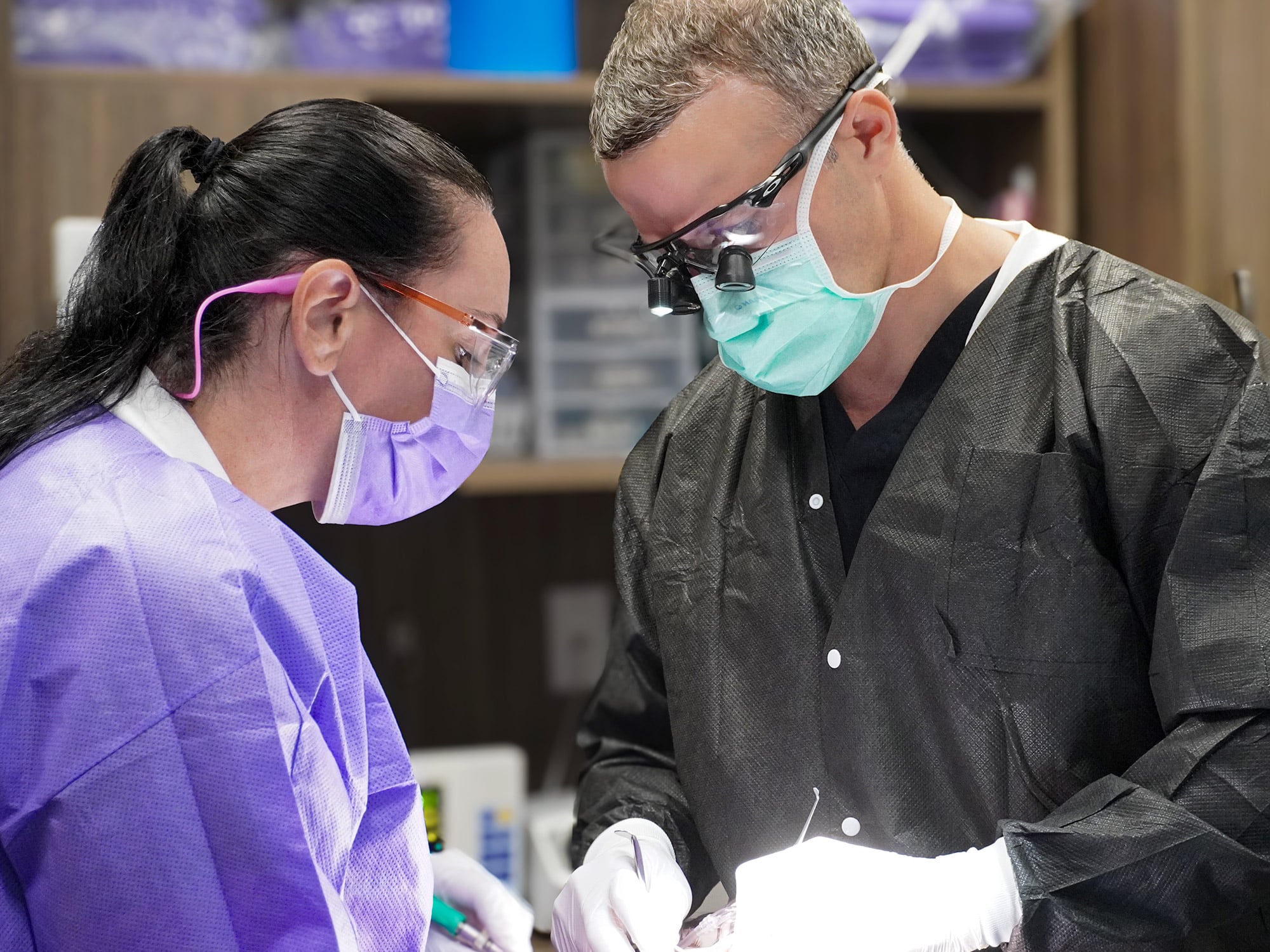 Two people in medical scrubs and masks perform a dental procedure, focusing intently with specialized eyewear in a clinical environment.