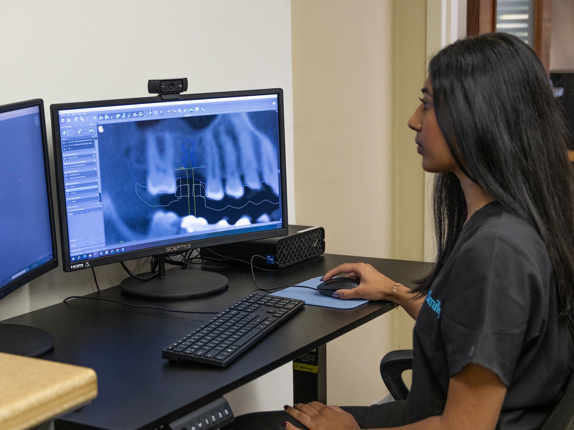 A person analyzes dental X-ray images on a computer, using dual monitors for precise examination. Technical software tools are visible on screen.