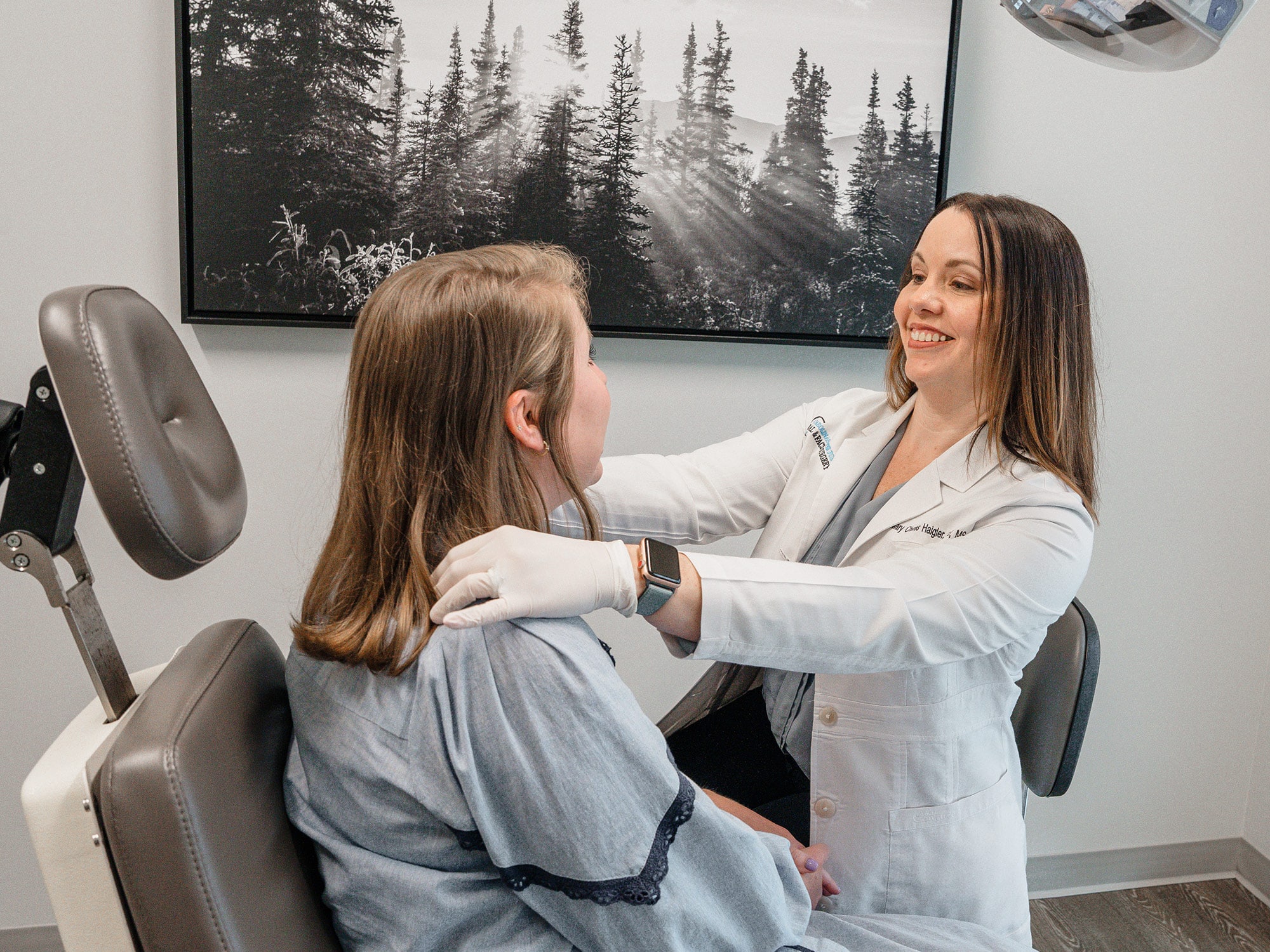 A person in a white coat assists another person in a clinic room with a nature photograph on the wall.