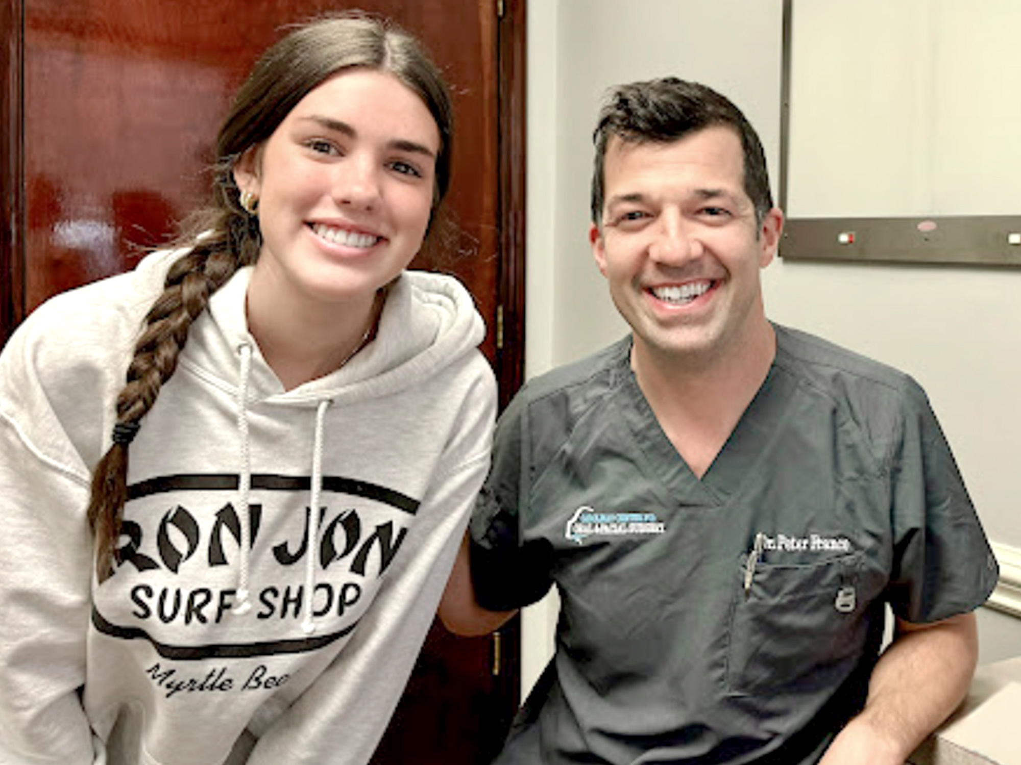 Two people smiling indoors; one wears a gray hoodie, the other in medical scrubs. They appear happy in what seems like an office setting.