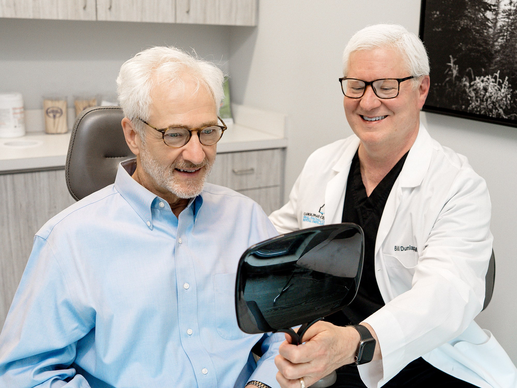 Two people in a medical office, one smiling and holding a mirror. Both are wearing glasses, with a white coat visible.