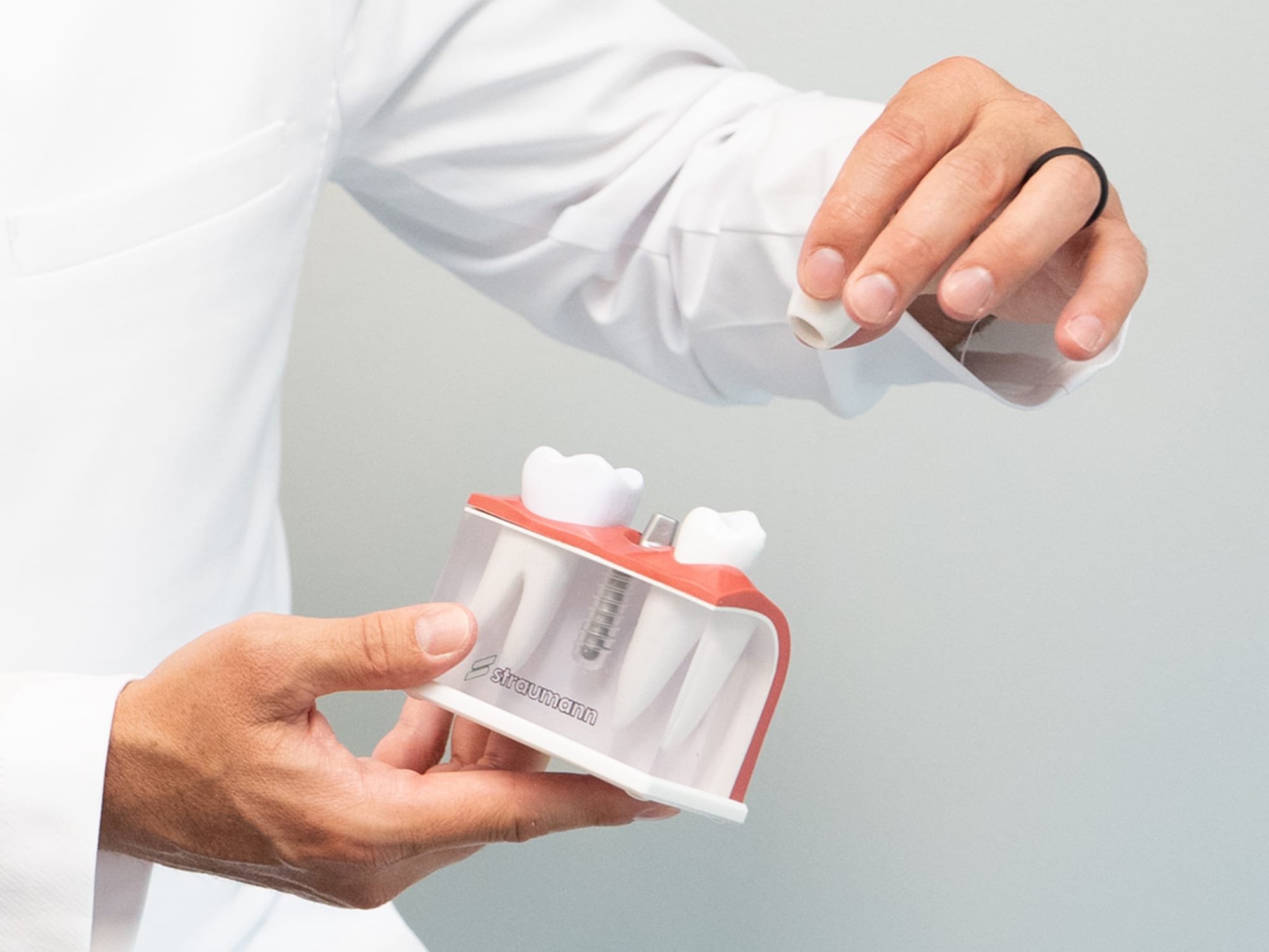 A person in a white coat holds a dental implant demonstration model, focusing on a removable artificial tooth component.