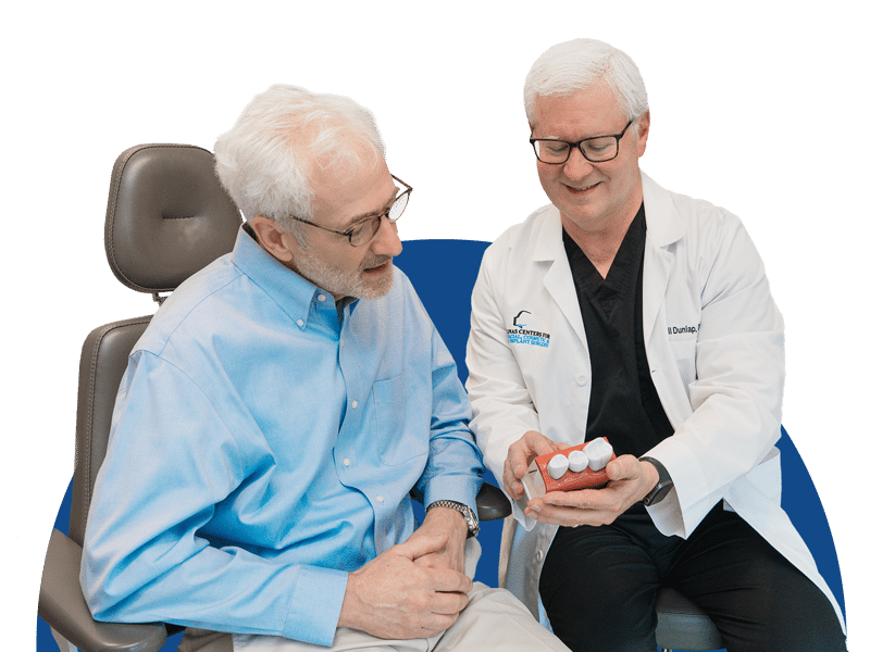 Two people sitting, one wearing a lab coat showing a dental model. They appear to be in a consultation setting.