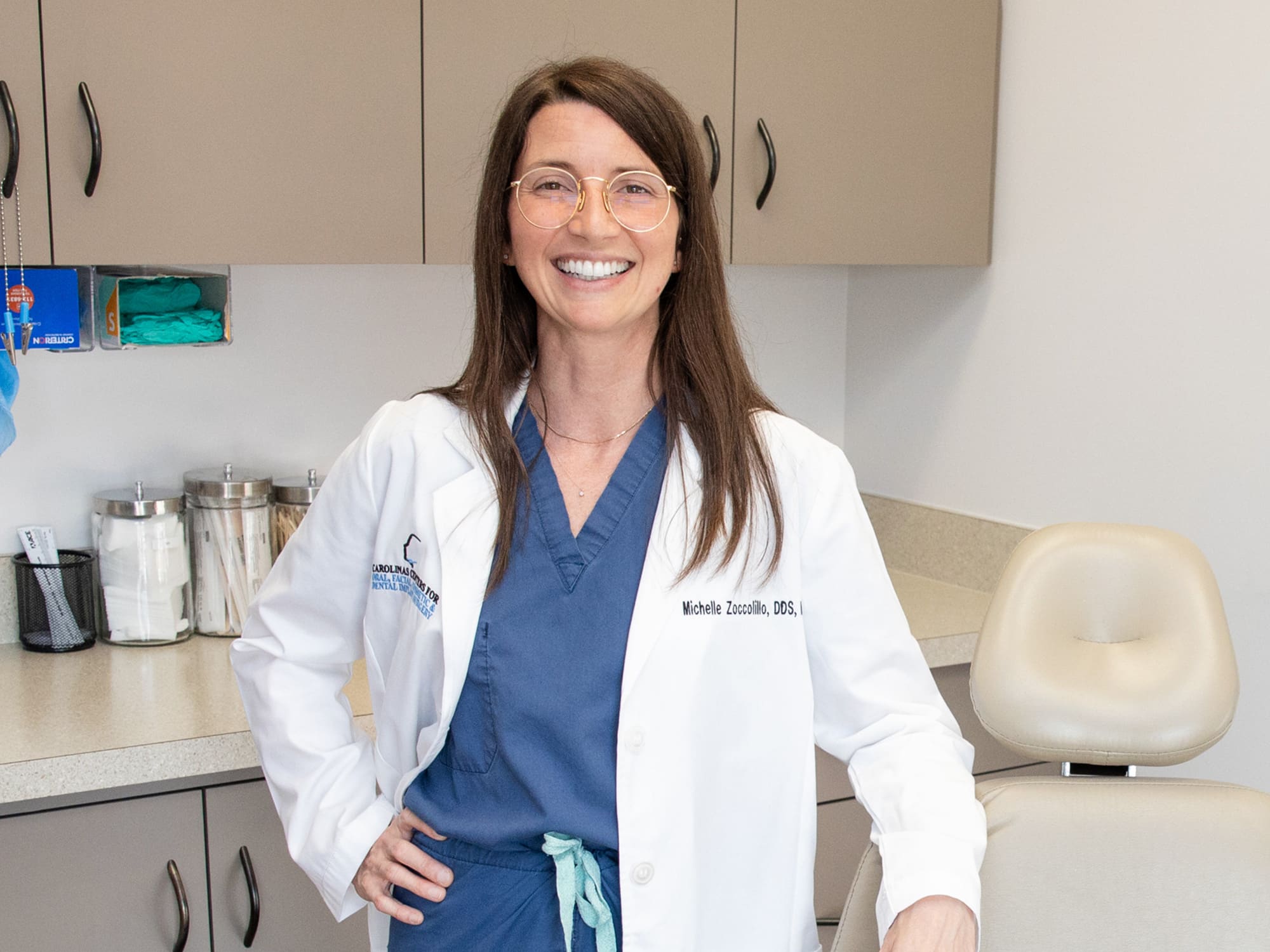 A person in a dental office smiles, wearing glasses and a white coat. Dental chair and supplies visible in the background.