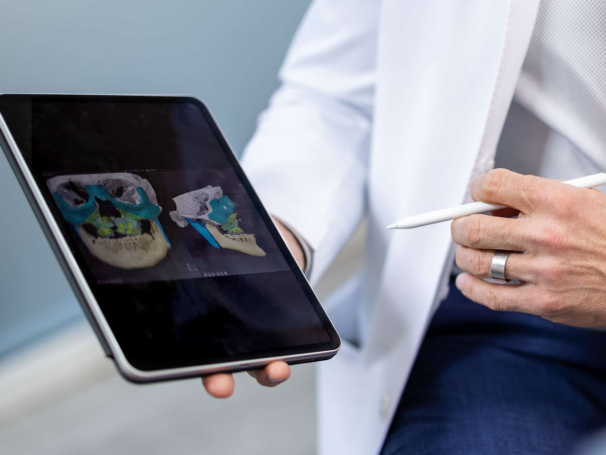 A person holds a tablet displaying 3D skull images, using a stylus. They wear a white coat, suggesting a medical or scientific context.