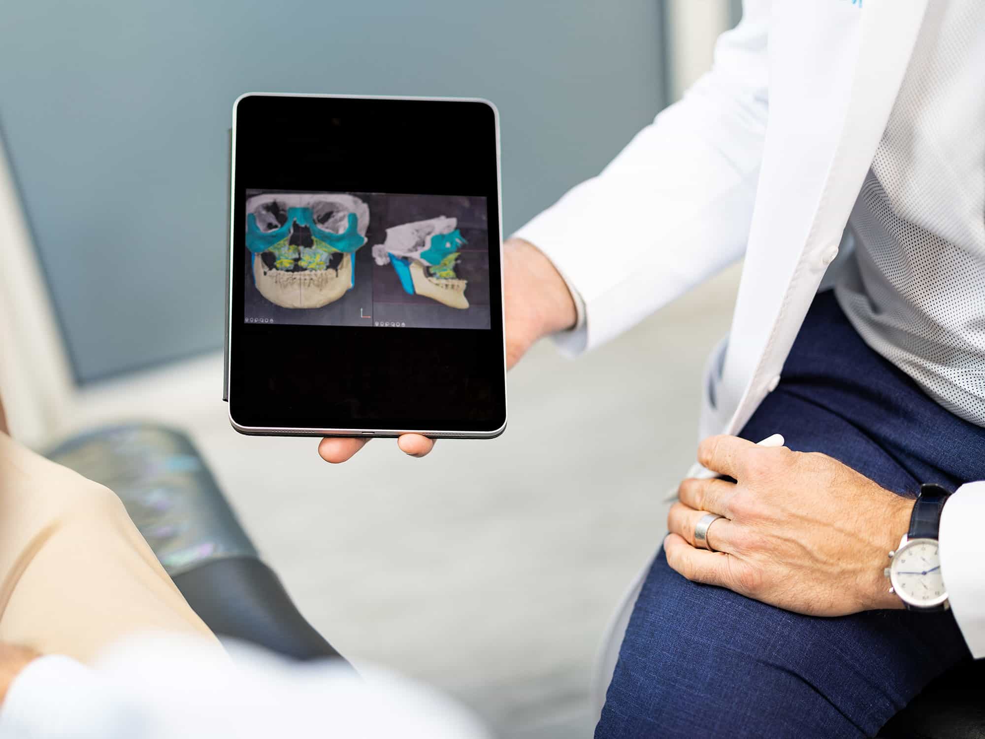 A person holding a tablet displaying 3D skull images, suggesting a medical or dental consultation, with focused hands and attire.