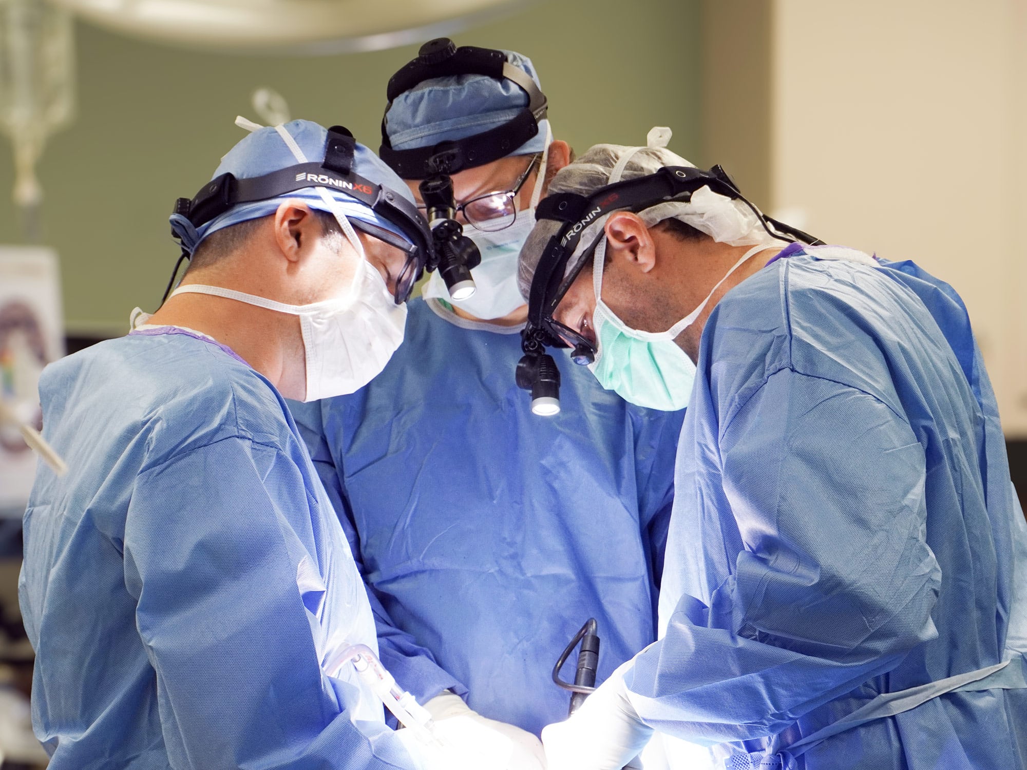 Three people in surgical attire focus intently on a task in a well-lit operating room, equipped with headlamps and sterile blue gowns.