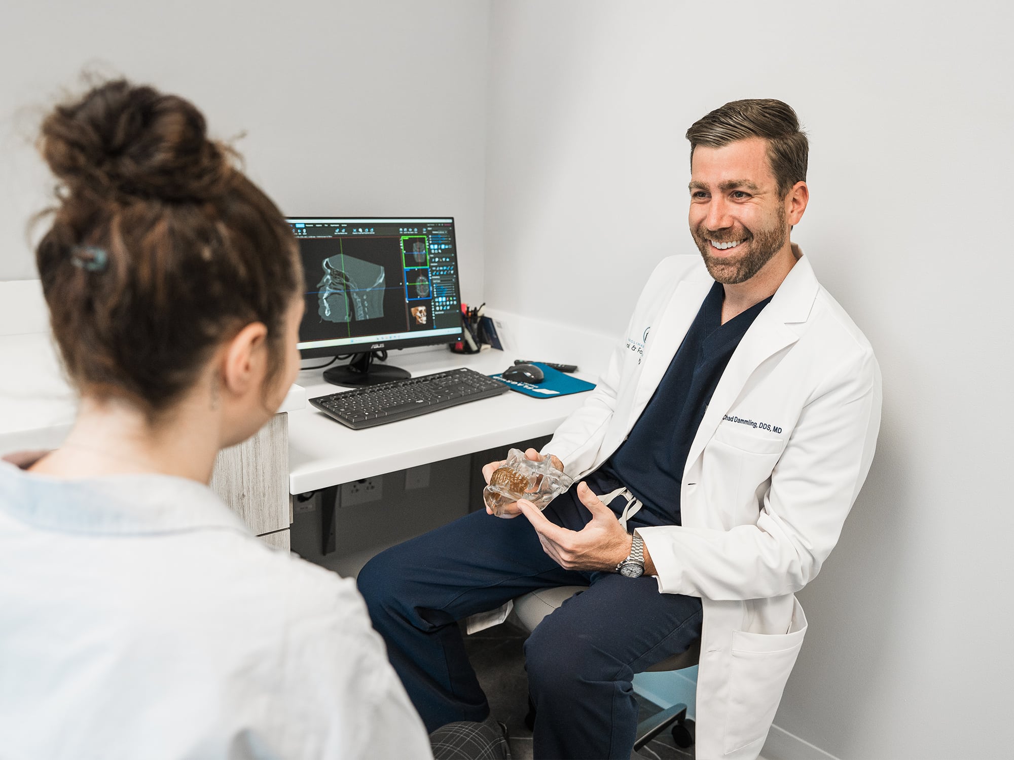 A person in a white coat discusses dental health with another person, showing a dental model. A computer screen displays dental images.