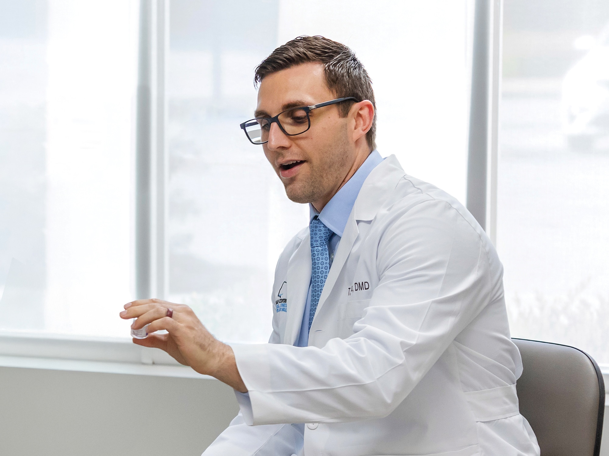 A person in a white coat with glasses gestures, sitting indoors, near a window, looking engaged, likely in a professional discussion.