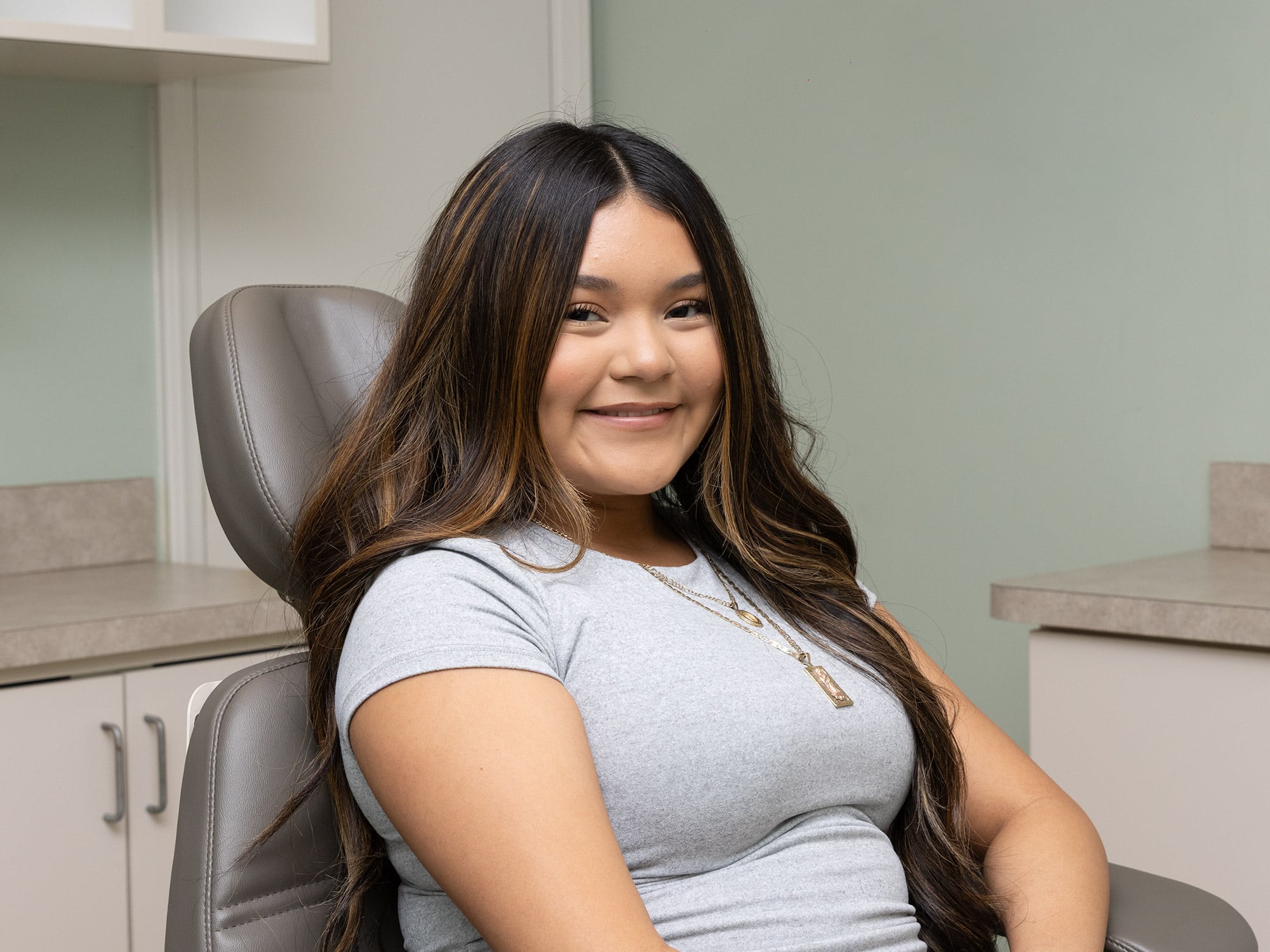A person sitting in a dental office chair, smiling. The background includes cabinets and a countertop, conveying a clean and professional setting.