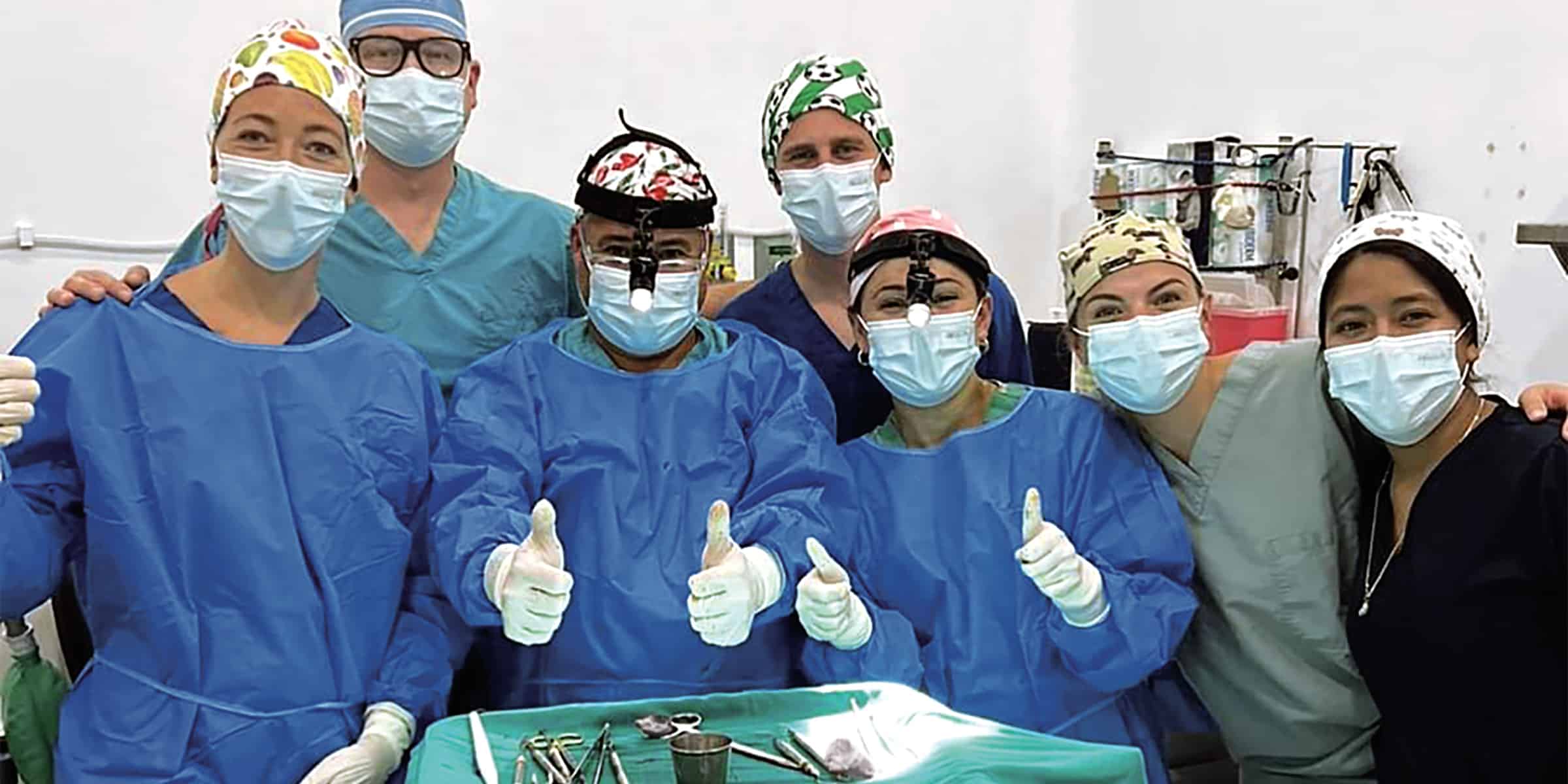 A group of healthcare professionals in scrubs and masks are smiling and giving thumbs up in a medical setting.