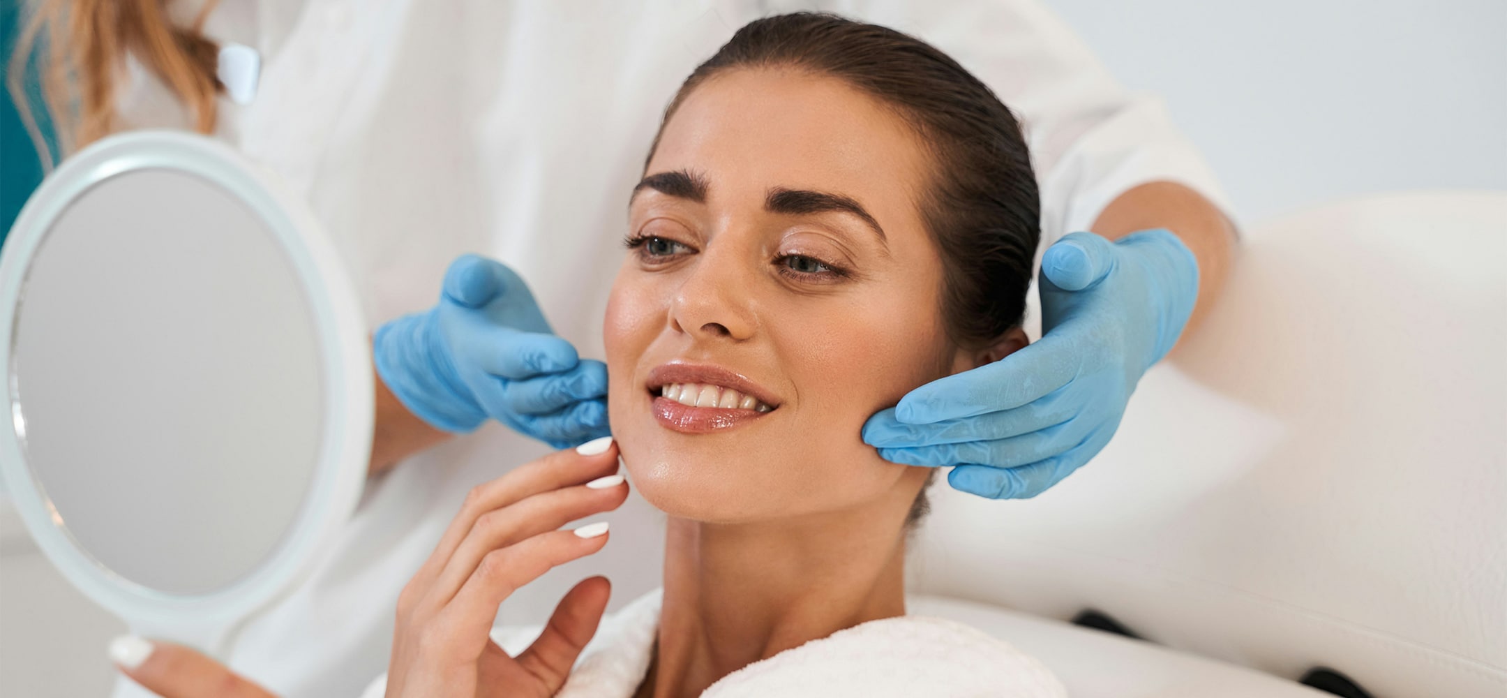 A person in a clinic holds a mirror, smiling as another person wearing blue gloves gently supports their face, suggesting a skincare consultation.