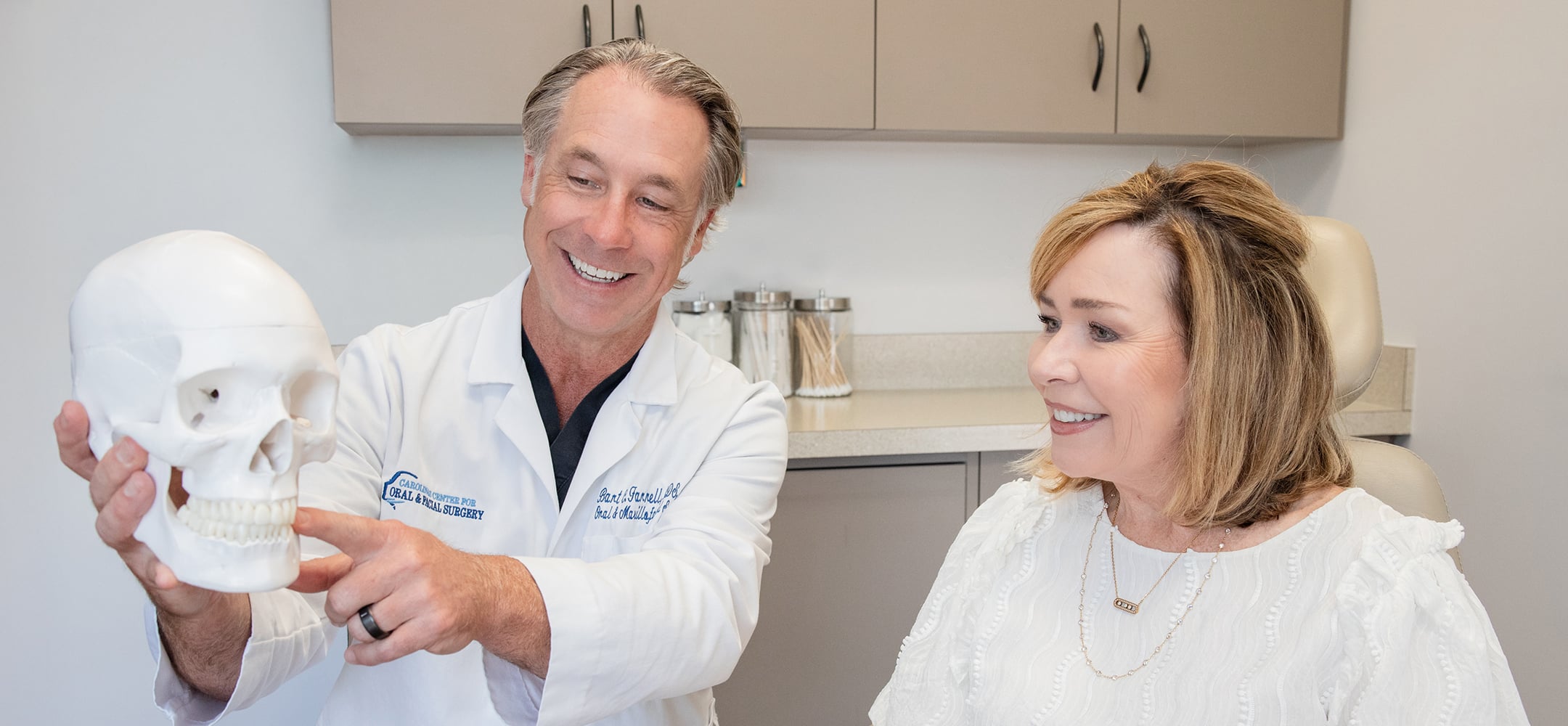 A person in a lab coat shows a model skull to another person, both smiling in a medical office.