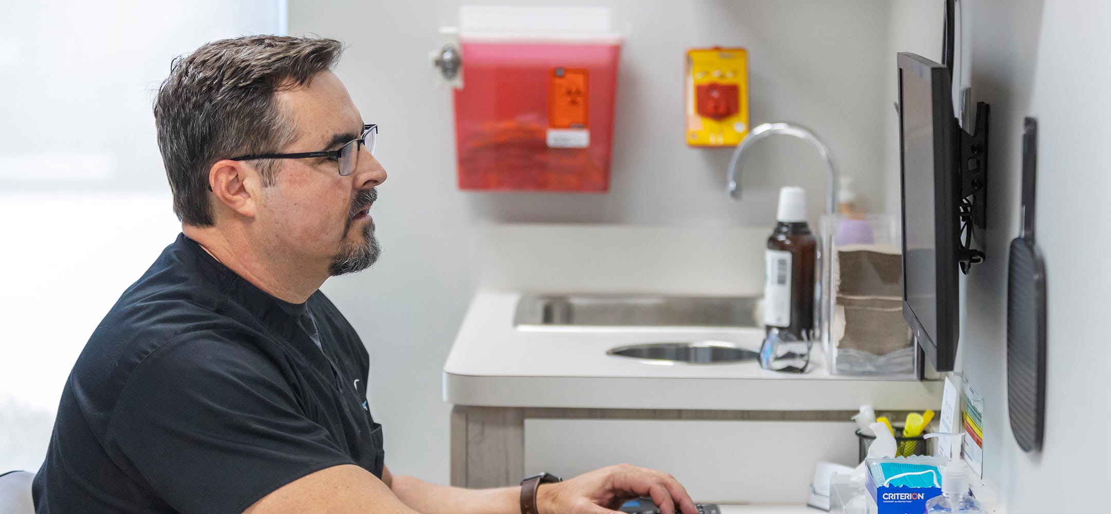 A person in medical attire works at a computer in a clinical setting, with medical supplies on the countertop and a sink nearby.