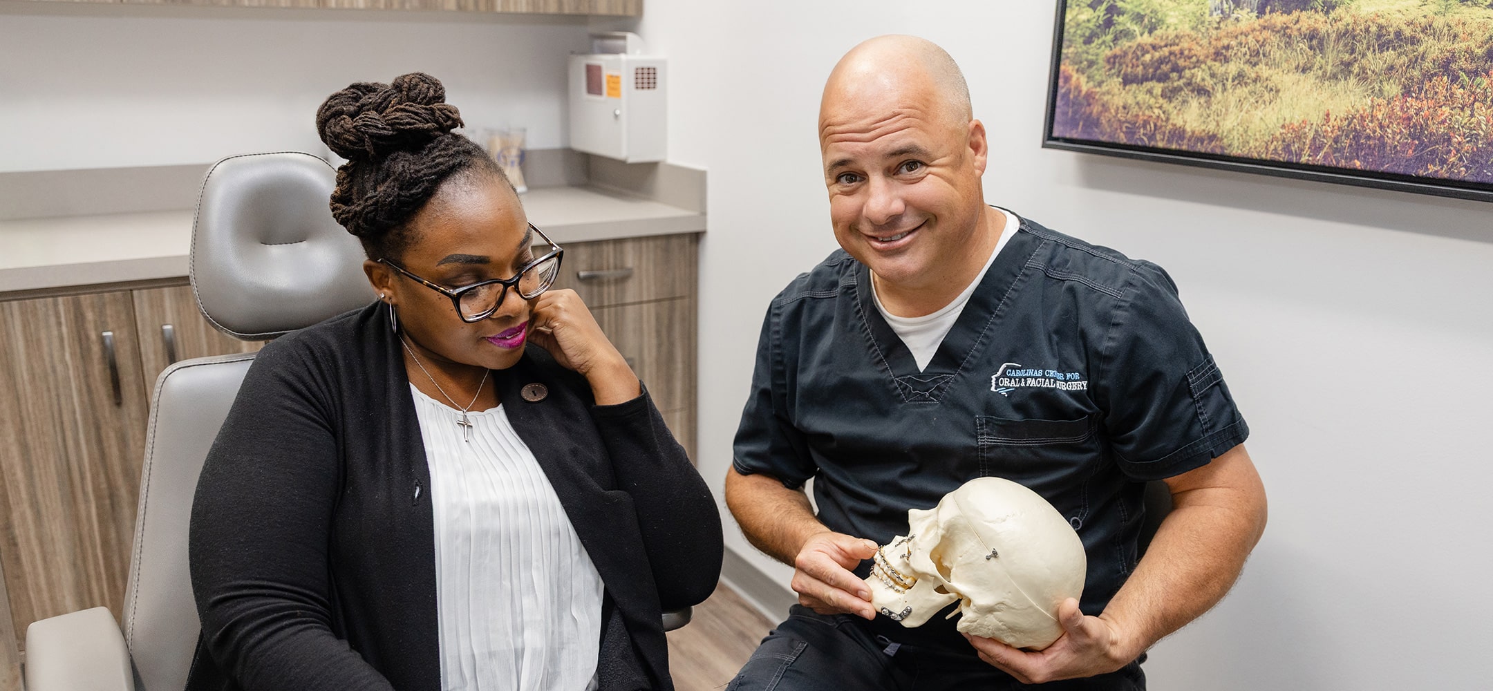 Two people in a medical office, one holding a skull model, discussing. Medical attire and office setting visible, no historical landmarks present.