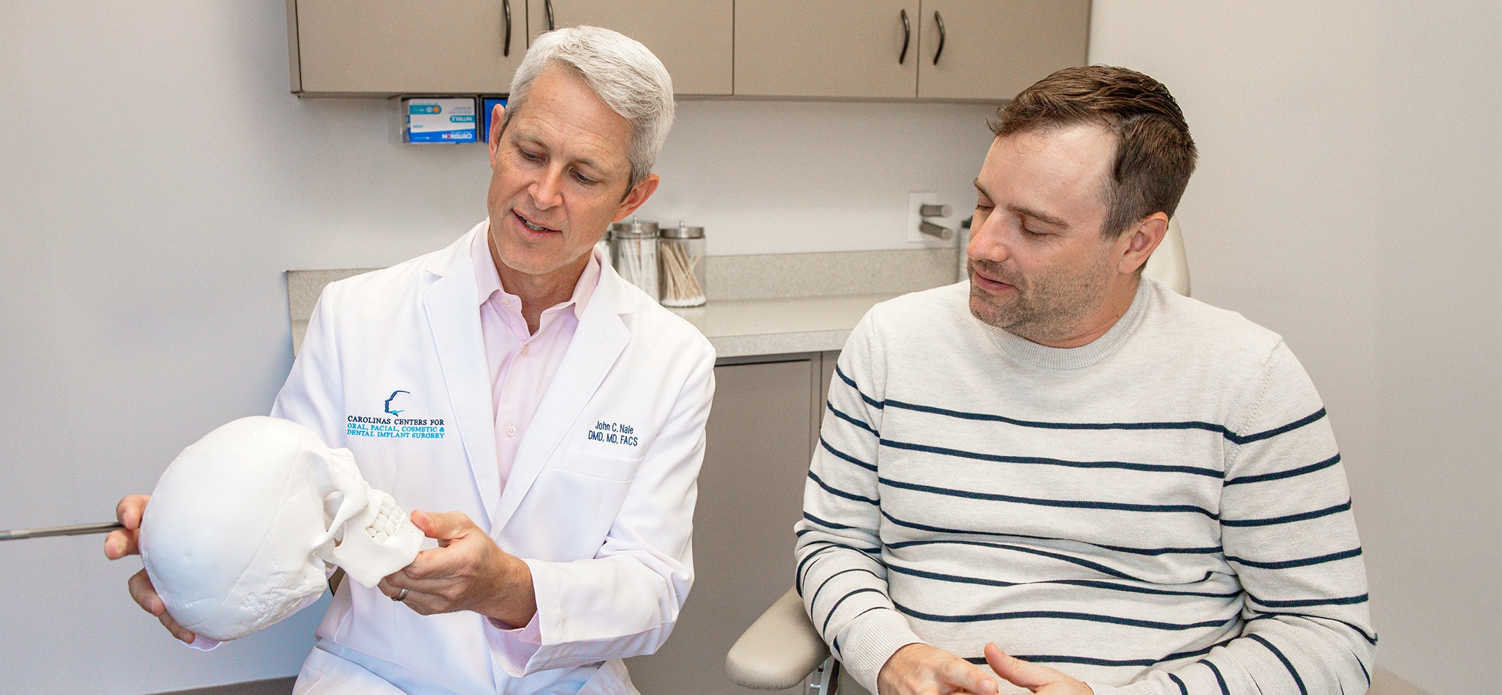A person in a lab coat shows a skull model to another person in a medical office setting.