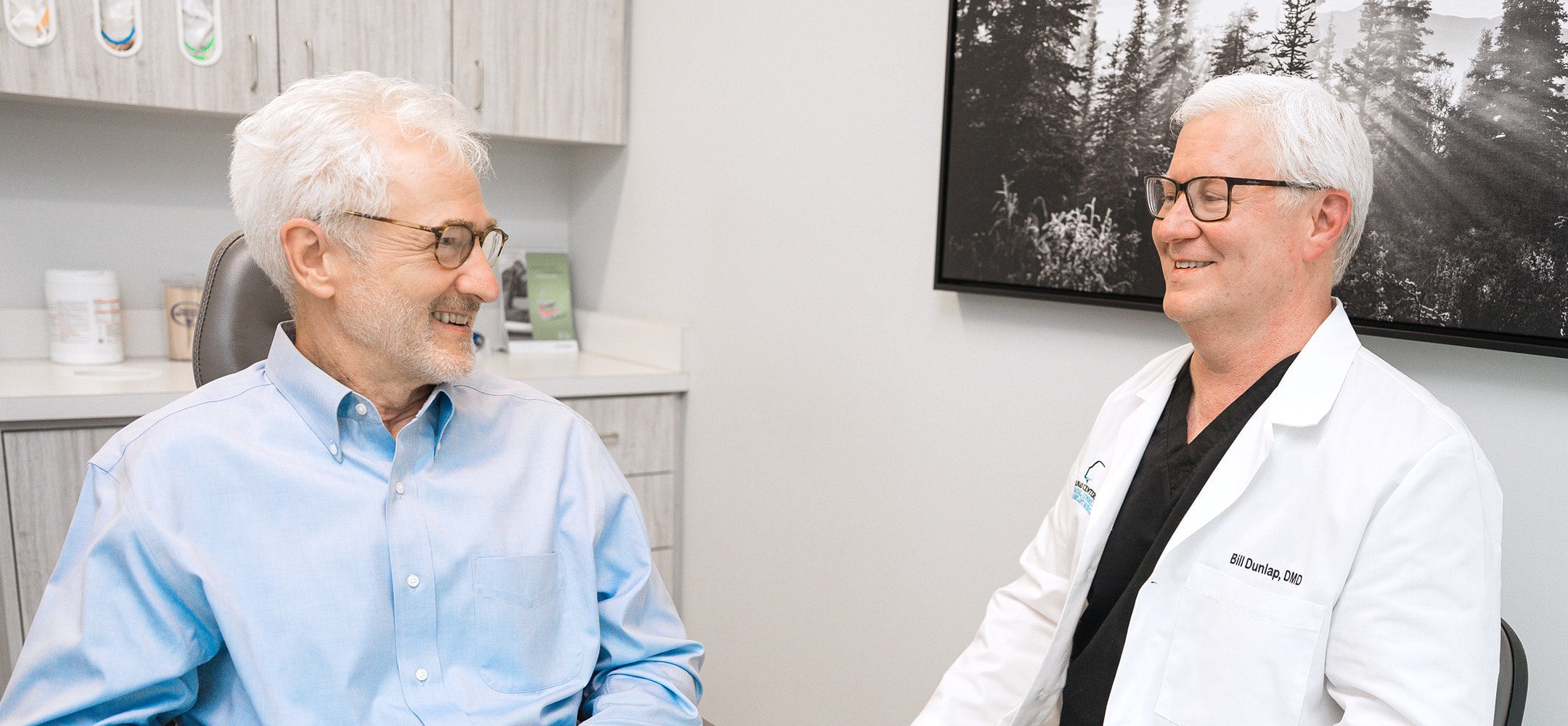 Two people smiling and talking in a medical office, one wearing a white coat. A black-and-white forest photo hangs on the wall.