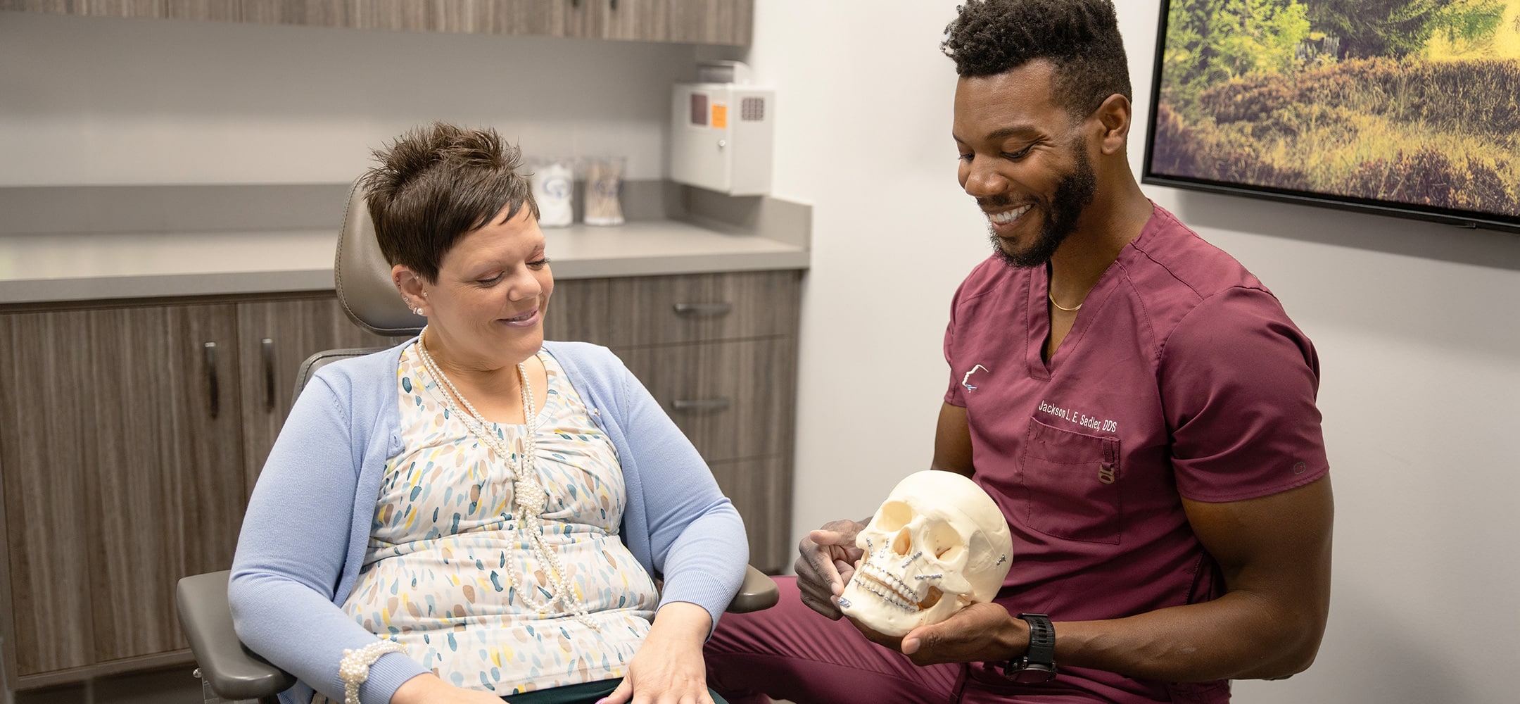 A person in scrubs shows a skull model to another person in an office, smiling and seated comfortably.