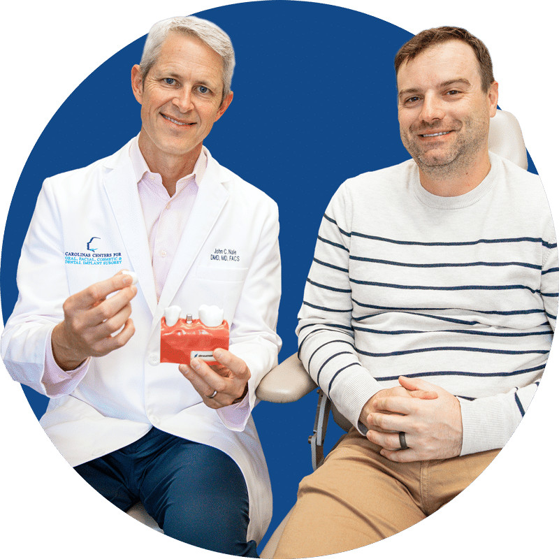 Two people are sitting against a blue background. One person in a white coat holds a dental model and an implant.