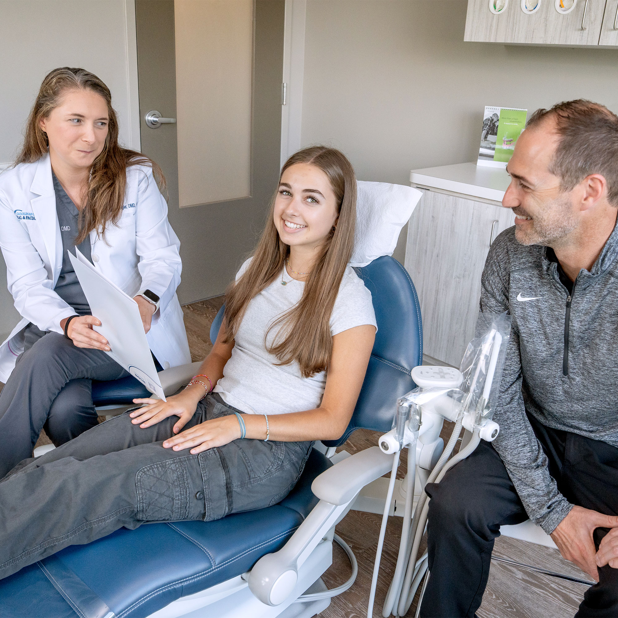 A person in a dental office converses with two others; one wears a white coat, and the patient smiles in a dental chair.