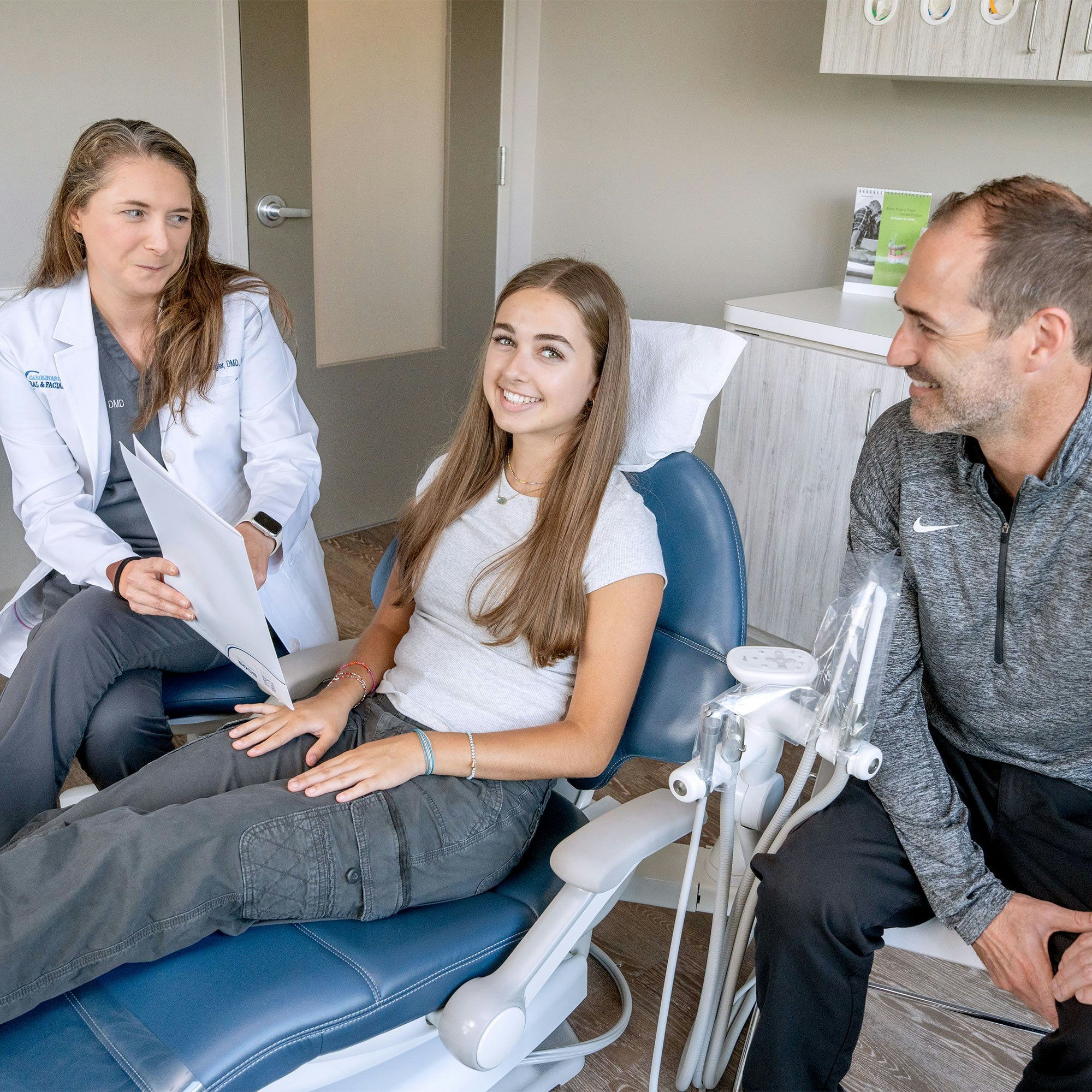 A person in a dental office converses with two others; one wears a white coat, and the patient smiles in a dental chair.
