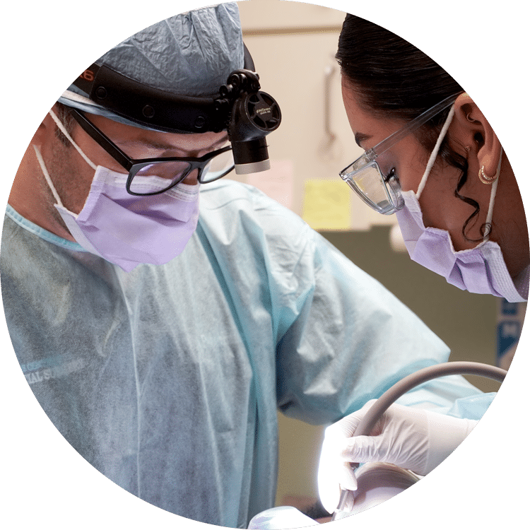 Two medical professionals in surgical attire focus intently, performing a procedure under bright lighting, emphasizing precision and teamwork in a clinical setting.