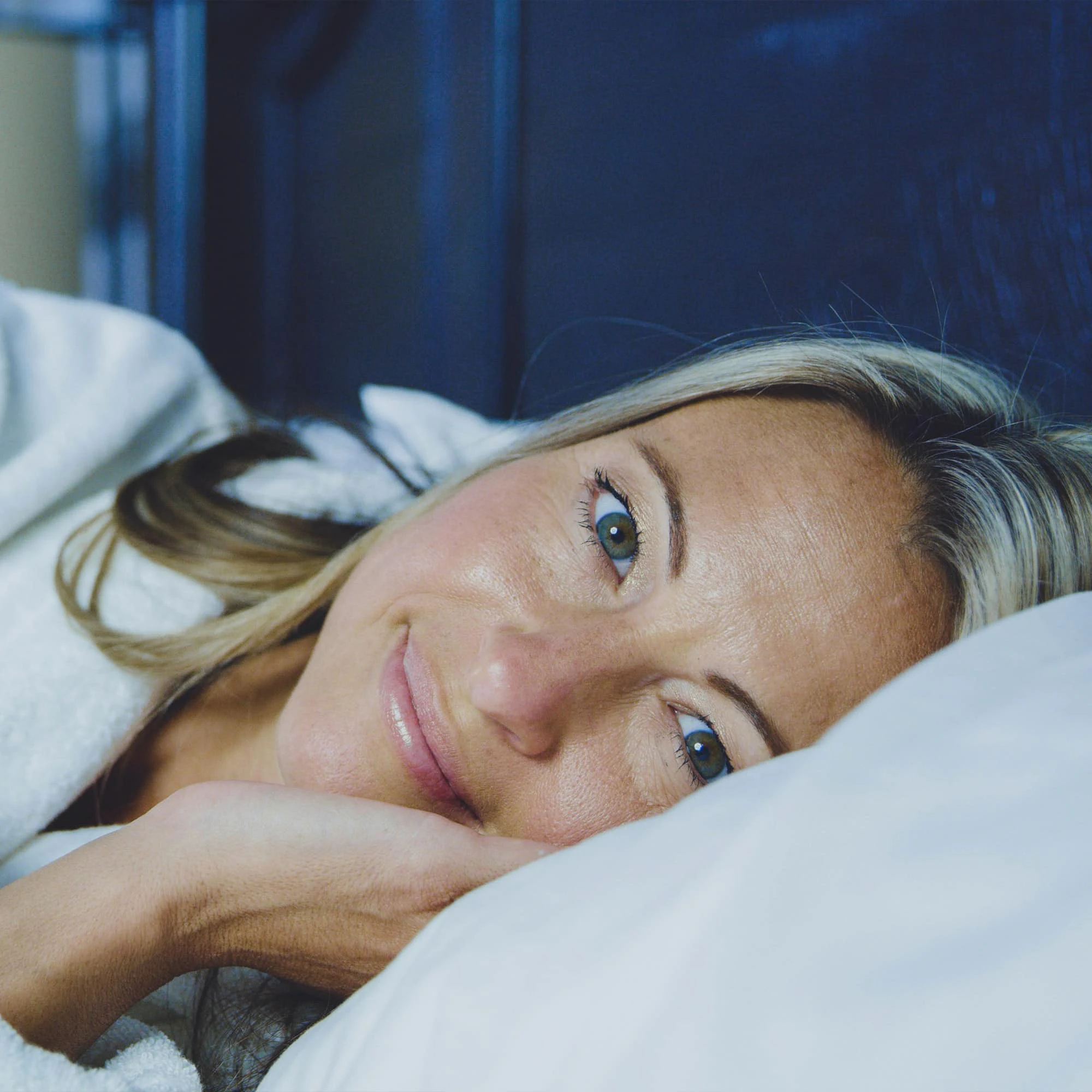 A person with blonde hair and blue eyes, wearing a white robe, lies comfortably on a bed, looking directly at the camera.