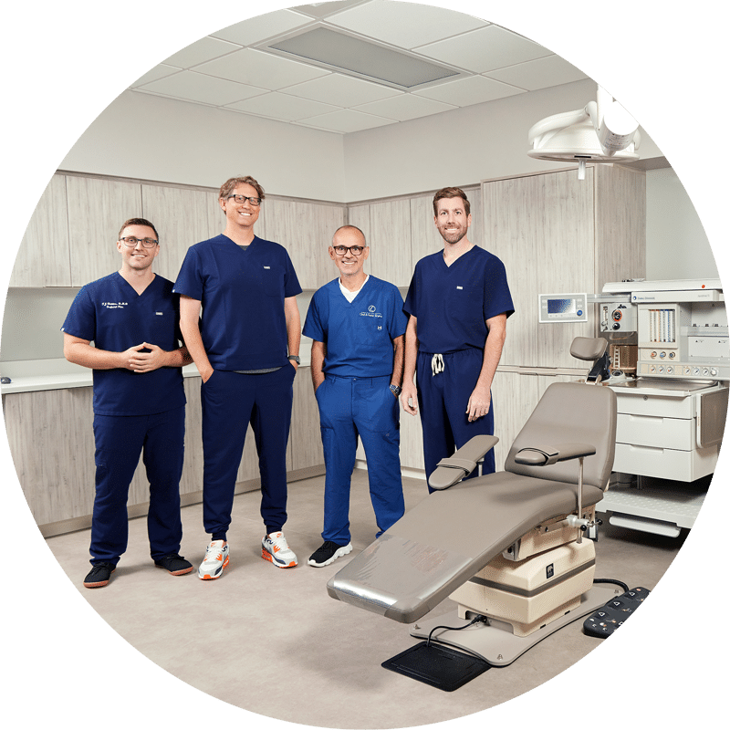 Four people in medical scrubs standing in a modern clinic room alongside an adjustable chair and medical equipment.