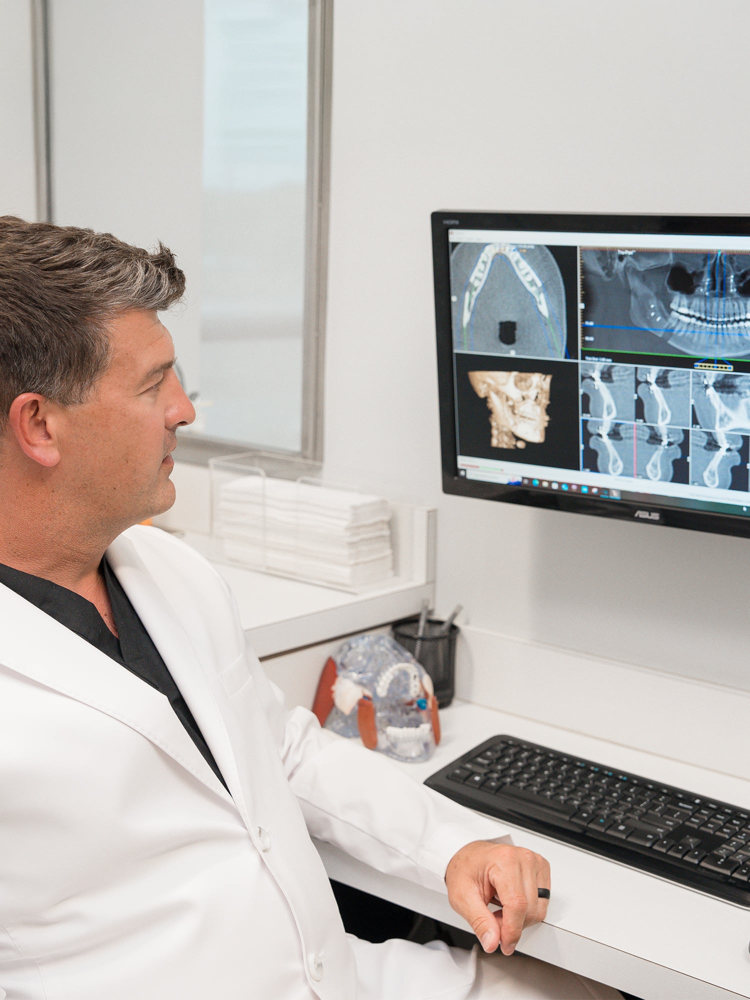 A person in a white coat observes dental X-rays on a computer screen in a clinical setting with dental models nearby.