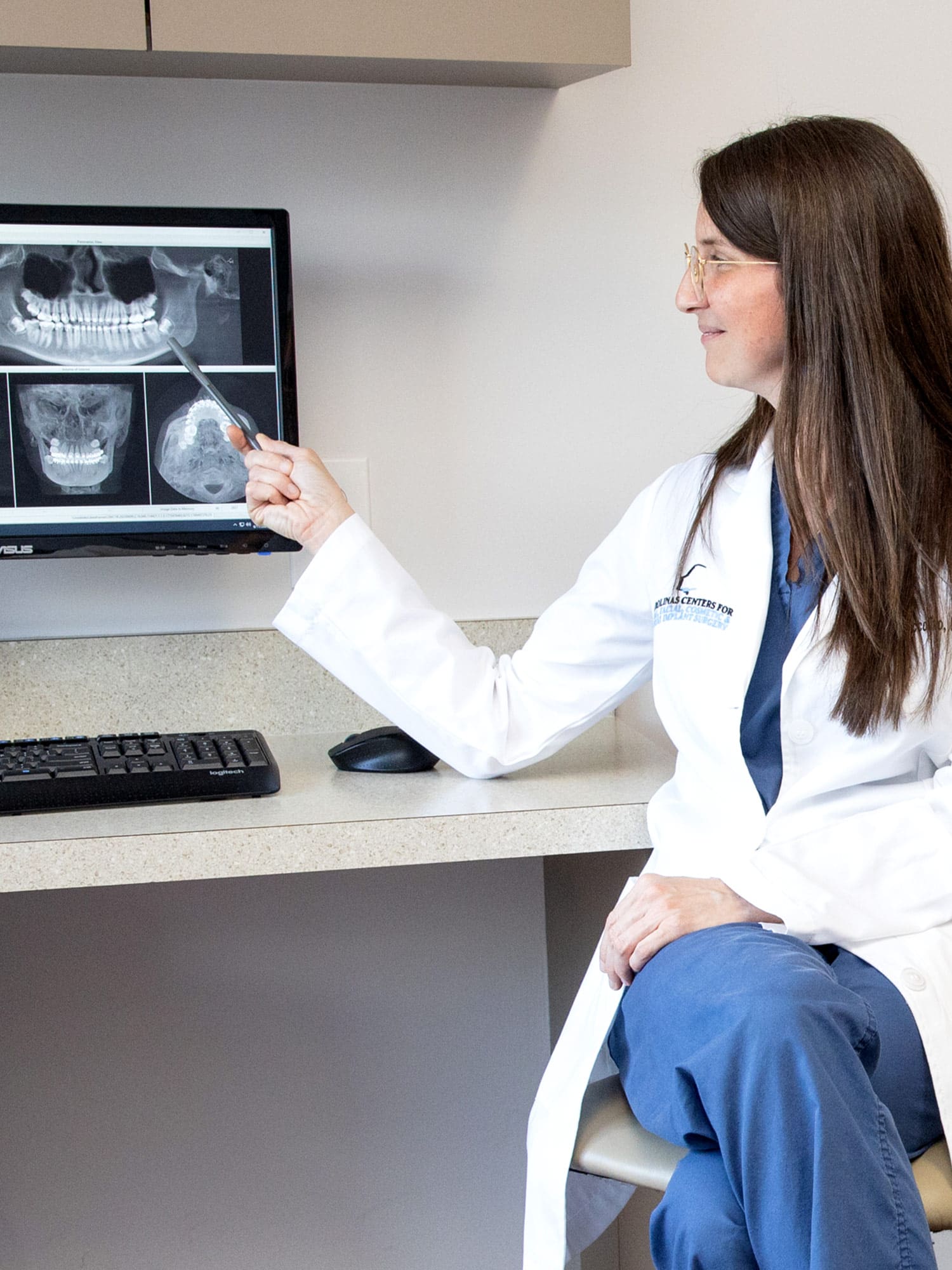 A person in a white coat points to dental X-rays on a computer screen, sitting in a clinical setting.