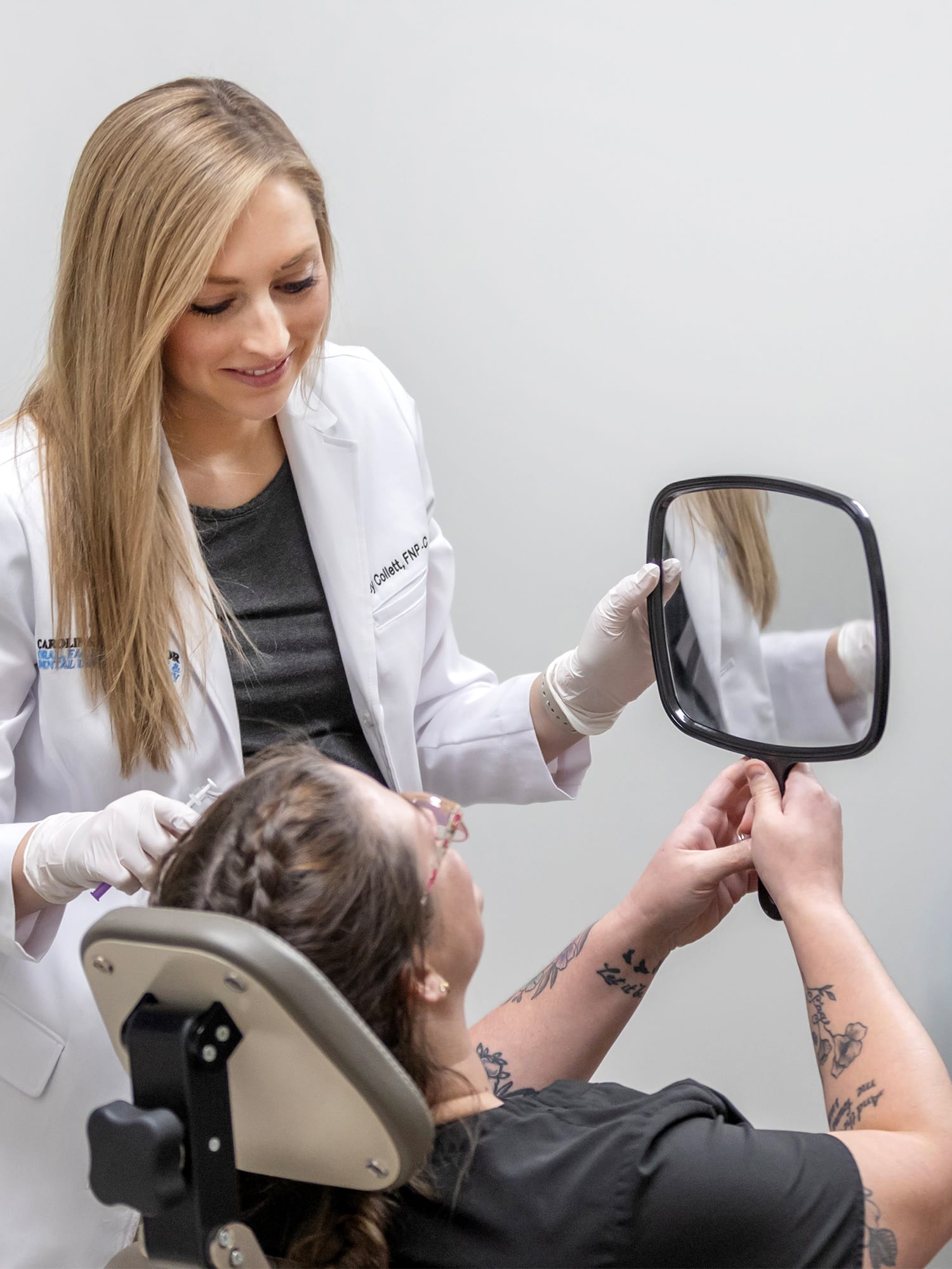 A person in a white coat helps another holding a mirror in a clinical setting, suggesting a consultation or medical procedure.