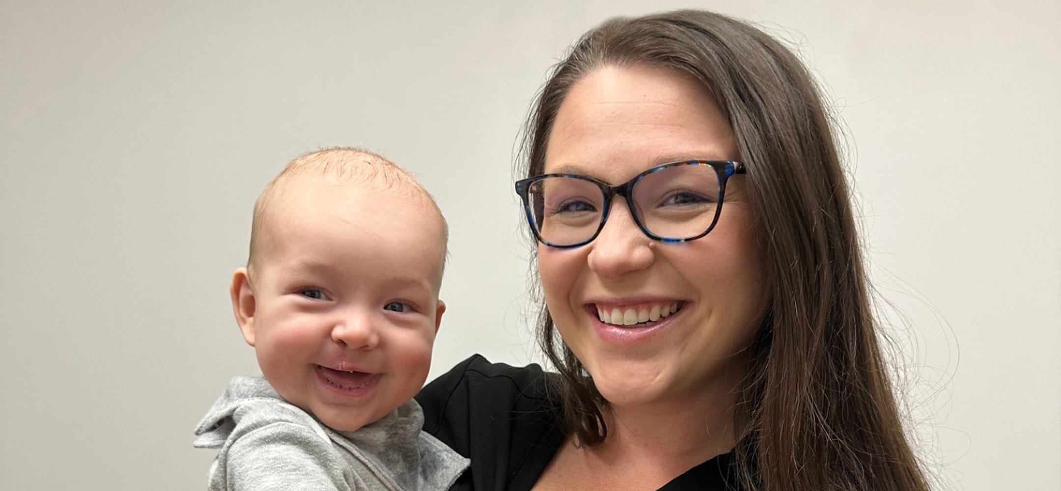 A smiling person with glasses holds a happy baby against a neutral background; both appear joyful and content.
