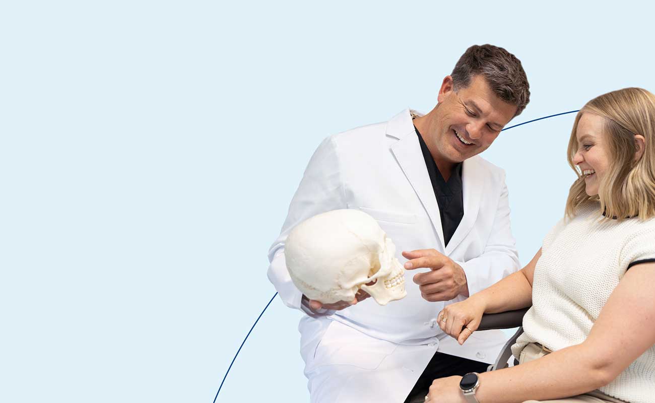Two people smiling, one wearing a white coat and holding a skull model, demonstrate dentistry or medical consultation on a plain background.