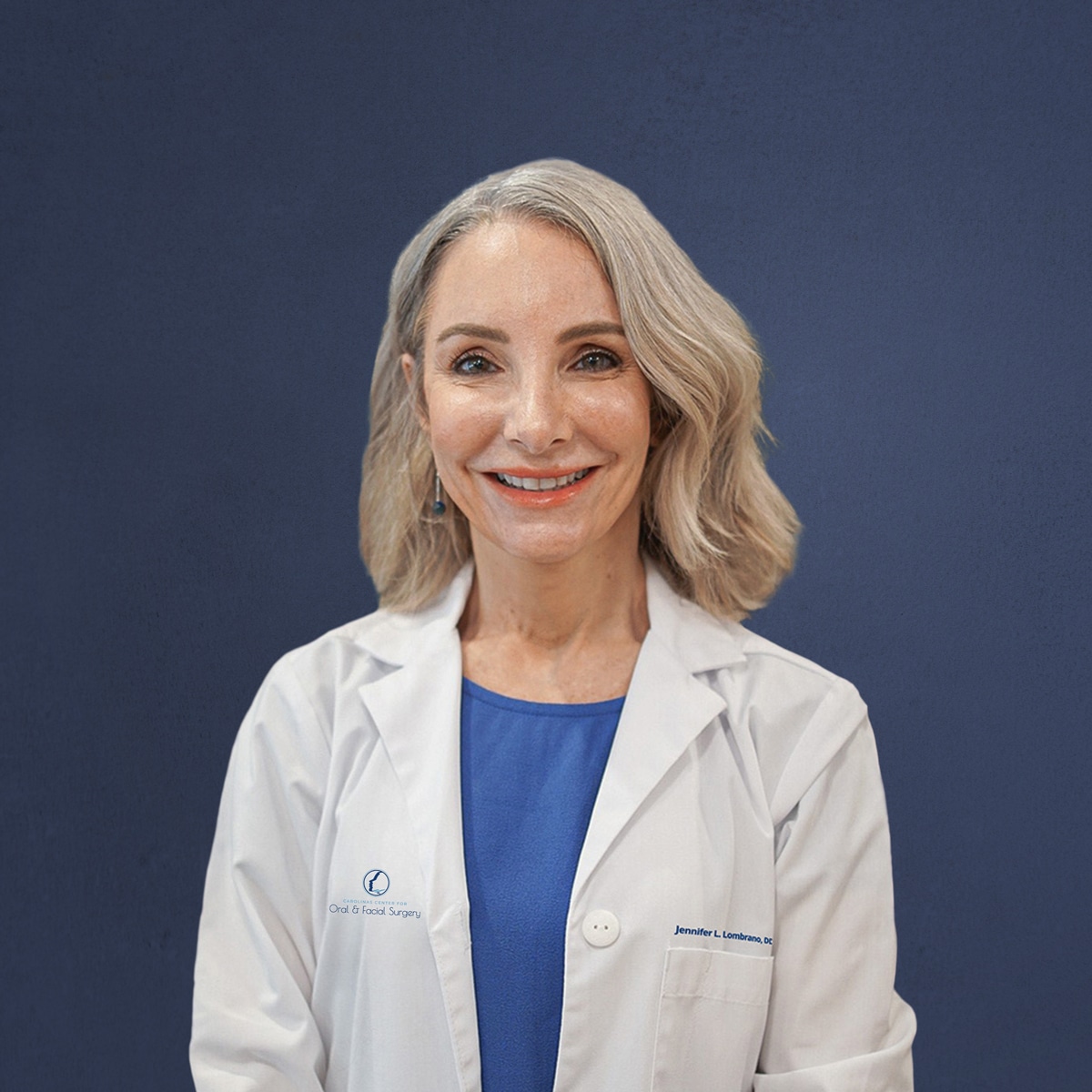 A person in a white coat smiles in front of a plain blue background, possibly in a professional setting.