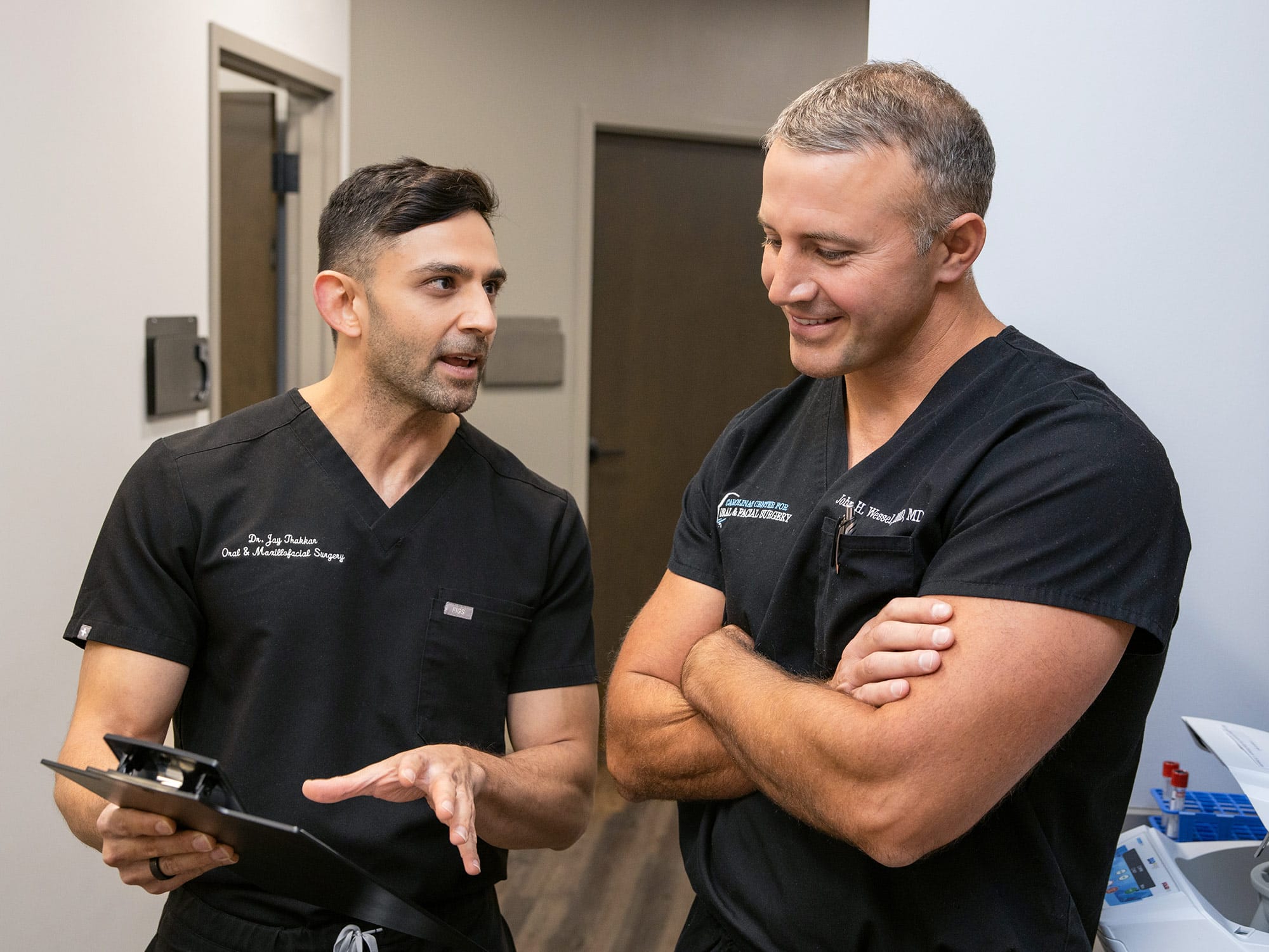 Two people in scrubs converse in a medical office, one holding a clipboard, standing near a white wall and a door.