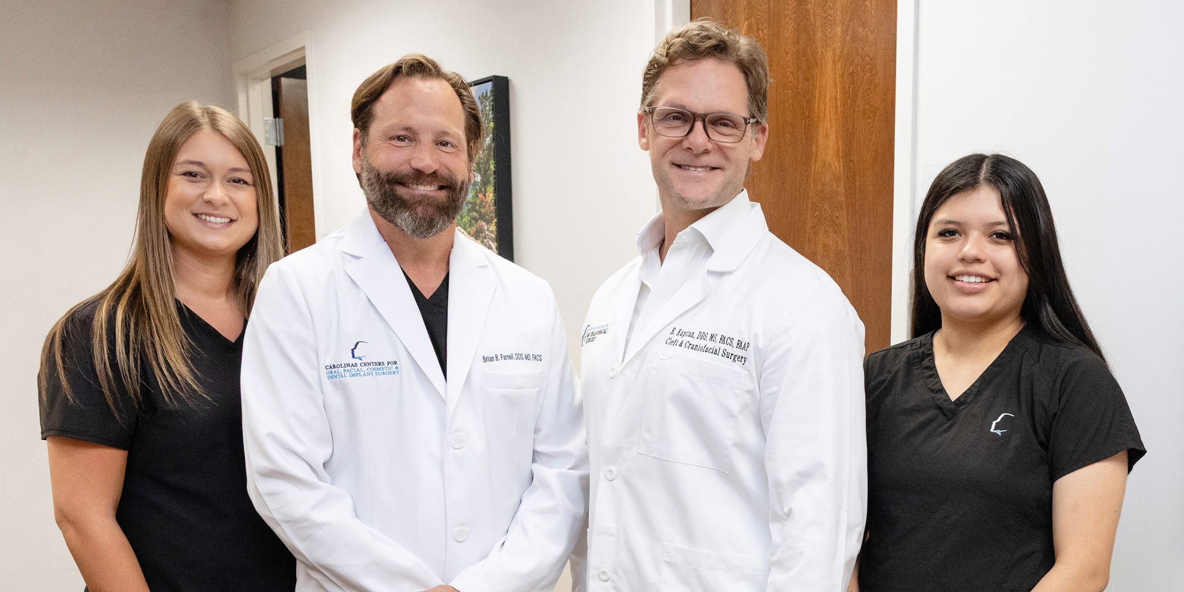 Four people in medical attire, two in white coats, two in black, standing in a clinical setting with a painting on the wall.