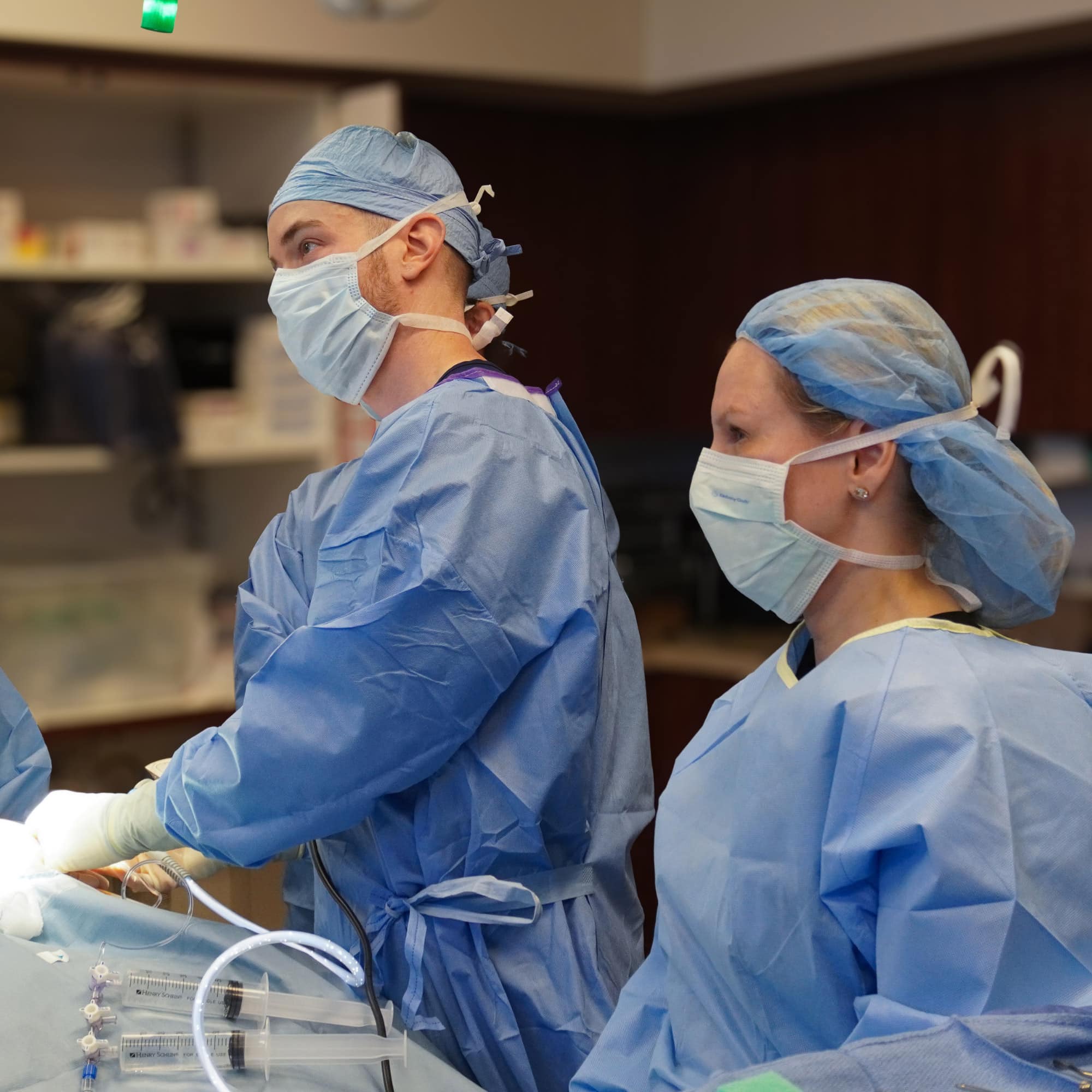 Medical professionals in an operating room, wearing blue surgical gowns and masks, focus intently on a procedure, with medical tools visible.