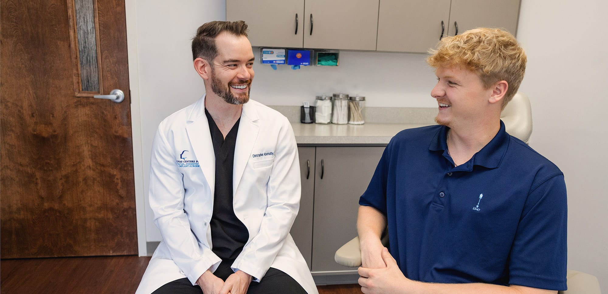 Two people sitting in a medical office, smiling at each other, one wearing a white coat, the other in a blue shirt.