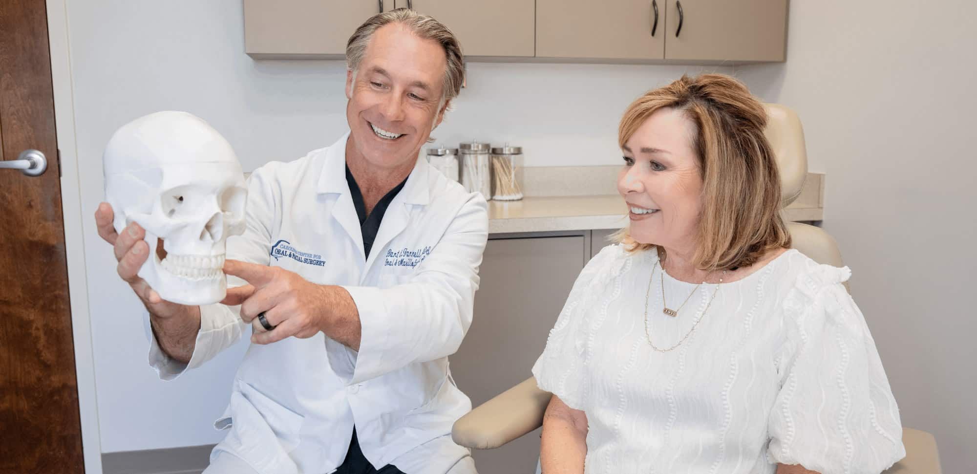 A person in a white coat shows a skull model to a smiling person in an office setting.