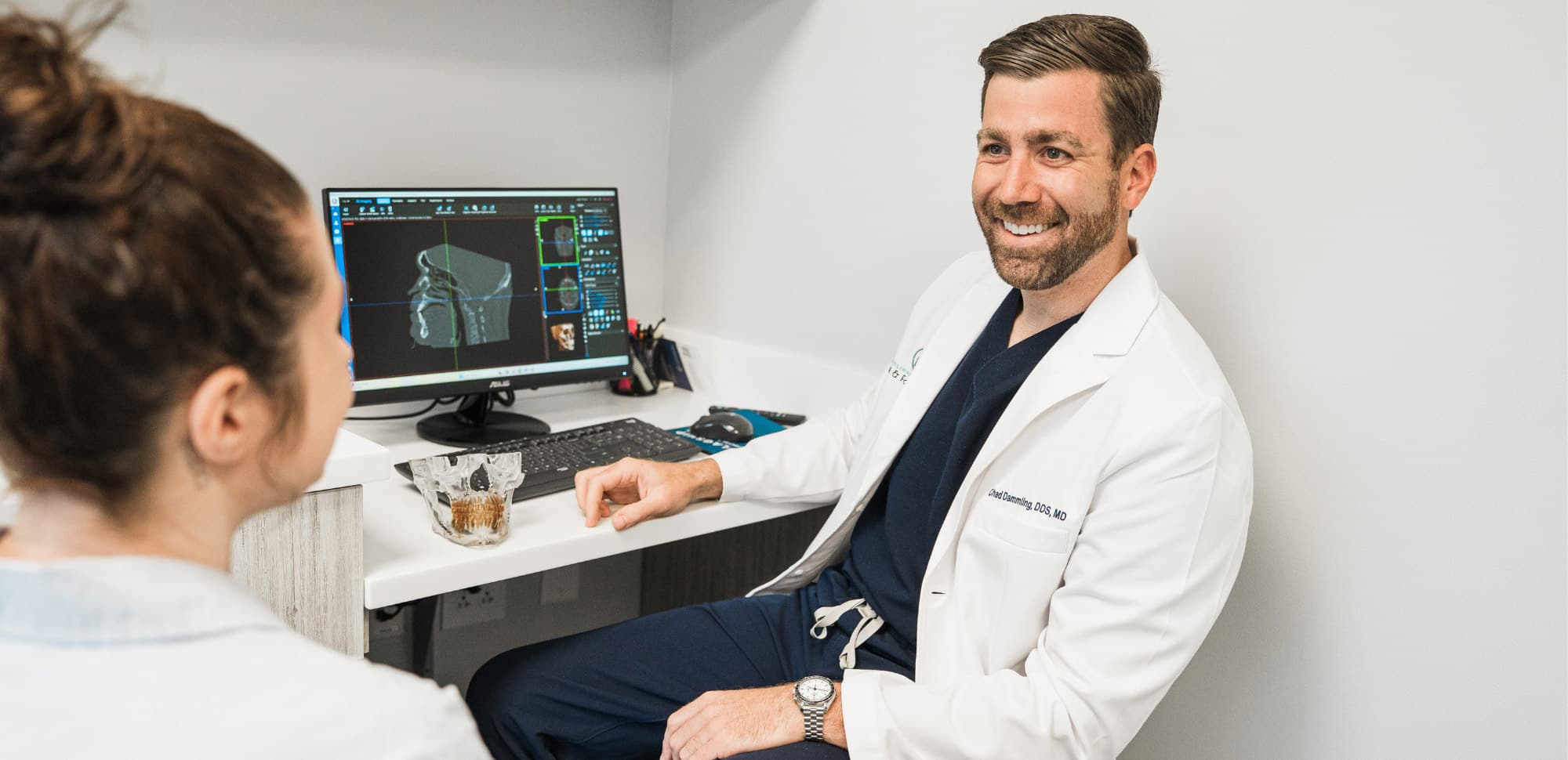 A person in a white coat smiling at another person in an office with a computer displaying a 3D image.