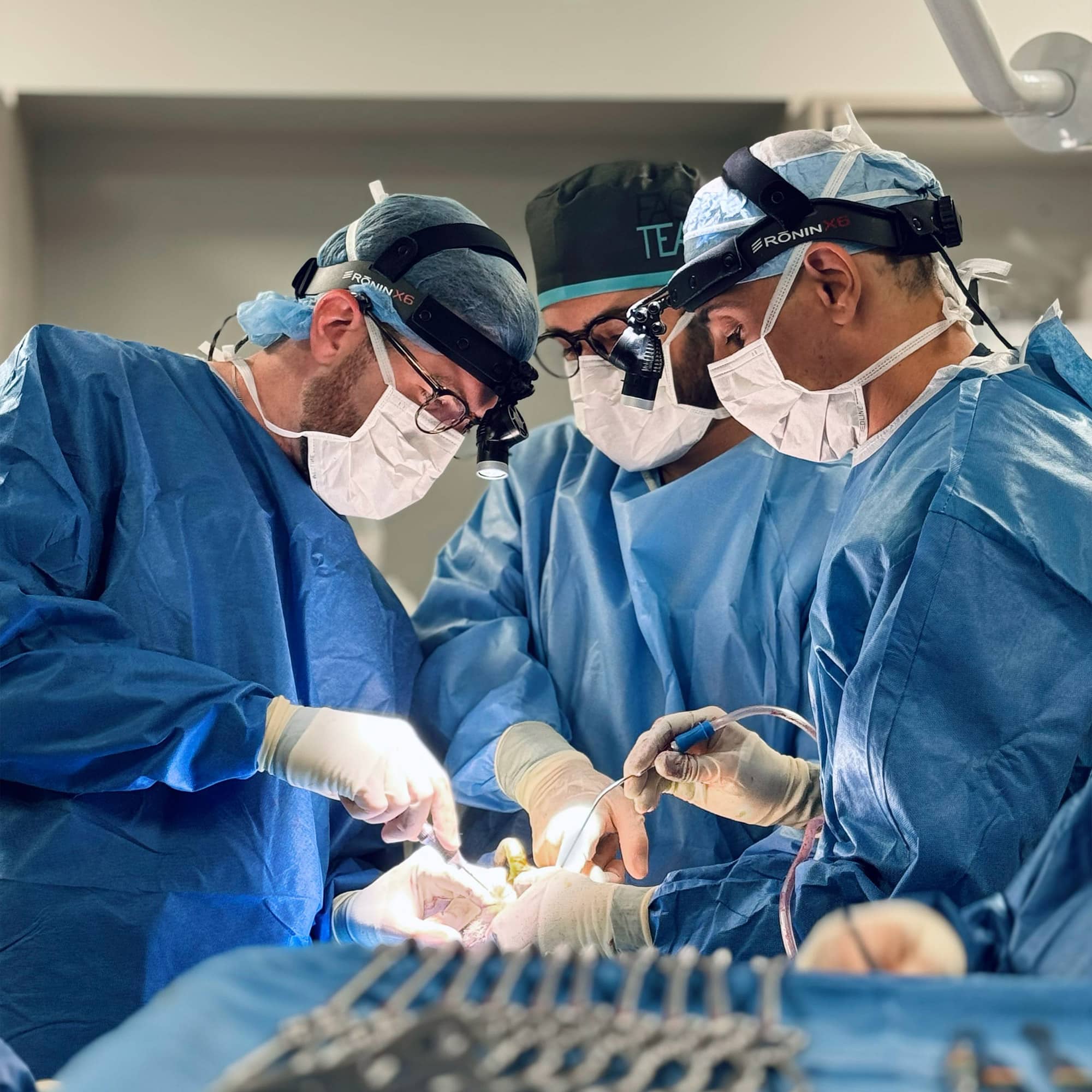 Three medical professionals in blue surgical attire perform surgery in a well-lit operating room, focusing intently on the patient.