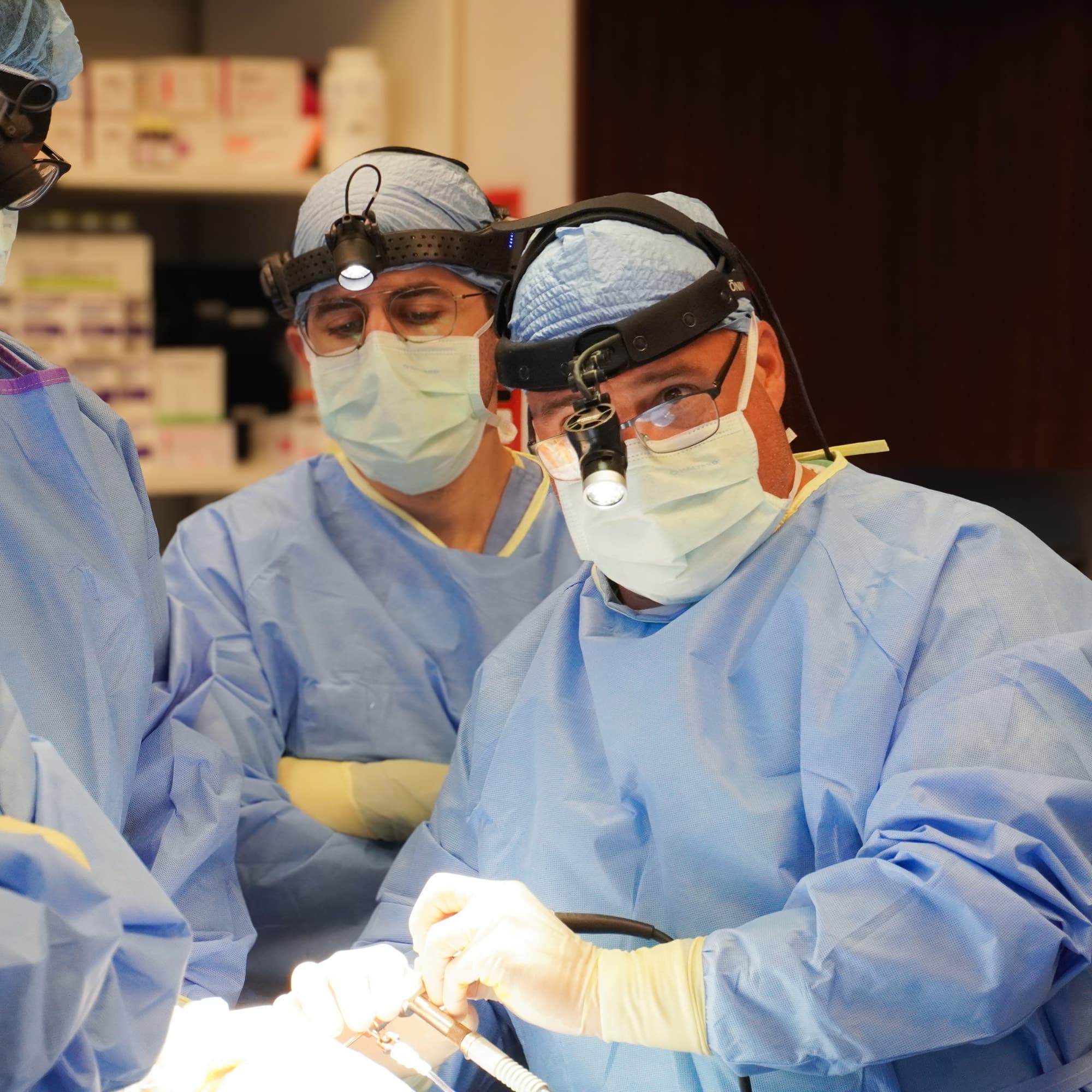 Three medical personnel in blue scrubs and masks perform surgery in a well-lit operating room, focused on the task at hand.