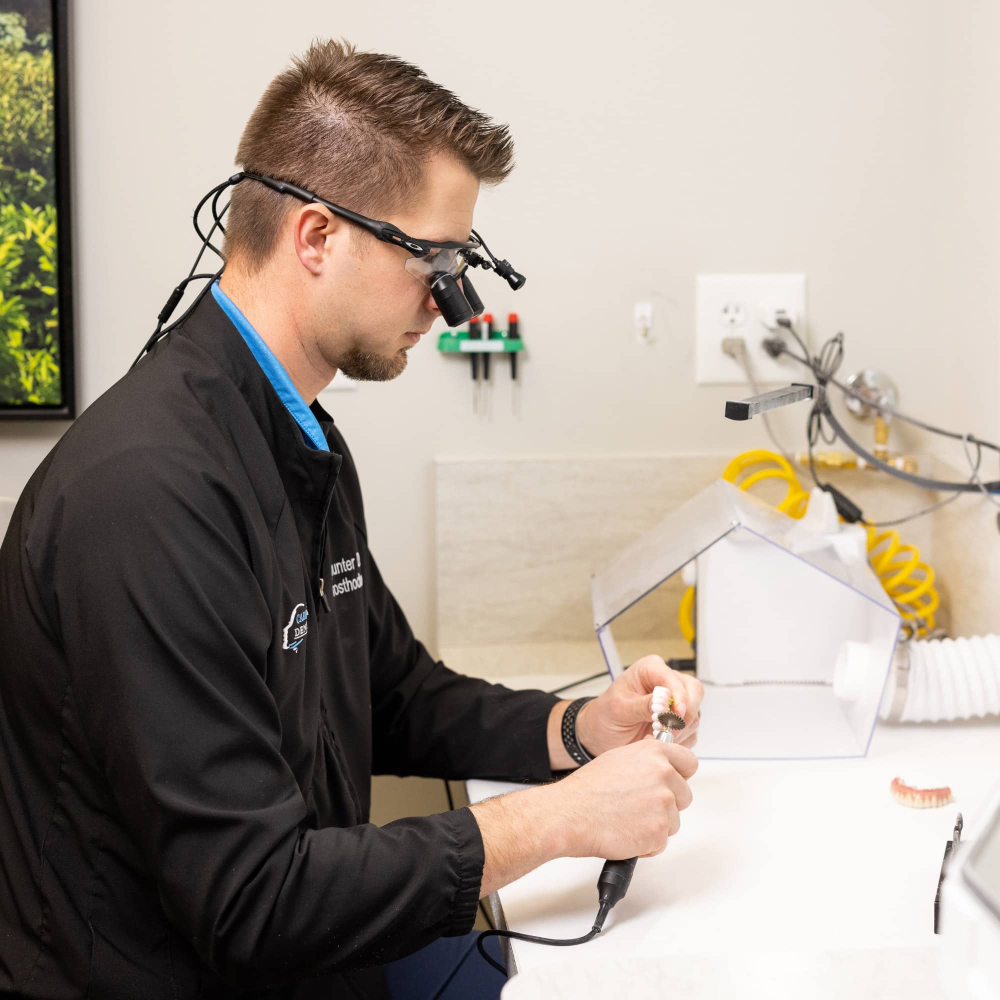 A person wearing magnifying loupes works meticulously on dental molds at a desk with dental equipment in a well-lit workspace.