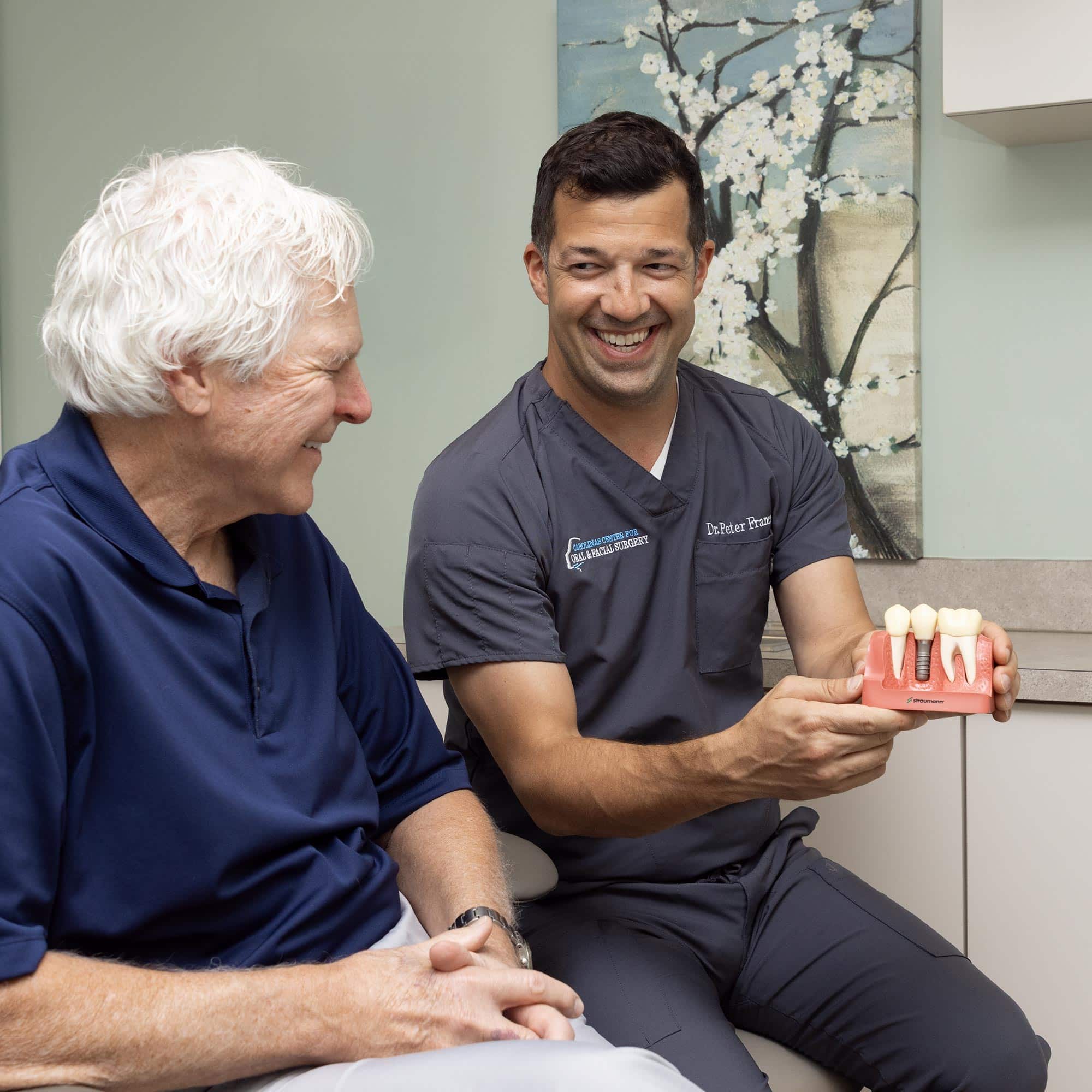 Two people in a dental office discuss a dental implant model. One person wears scrubs, and a floral painting decorates the background.